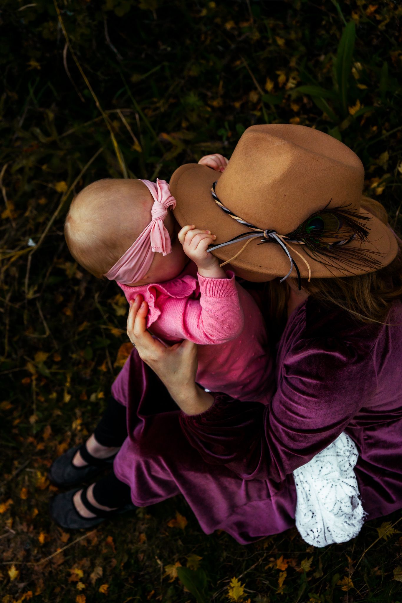 Overhead view of mother in burgundy dress and tan hat cradling baby in pink on autumn ground.