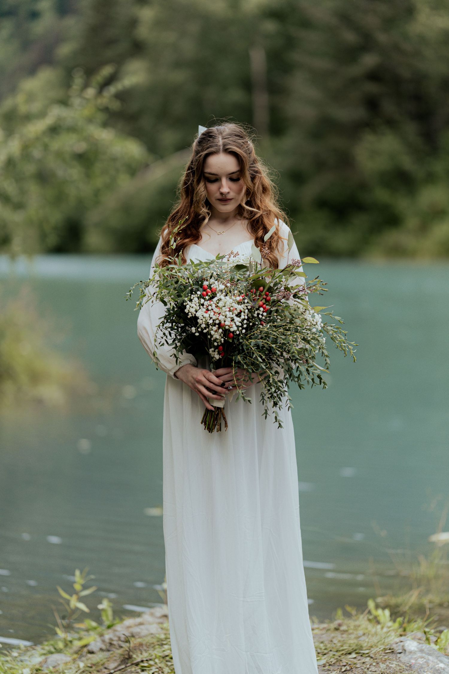 Bride in flowing white gown holds wildflower bouquet while standing beside a turquoise lake.