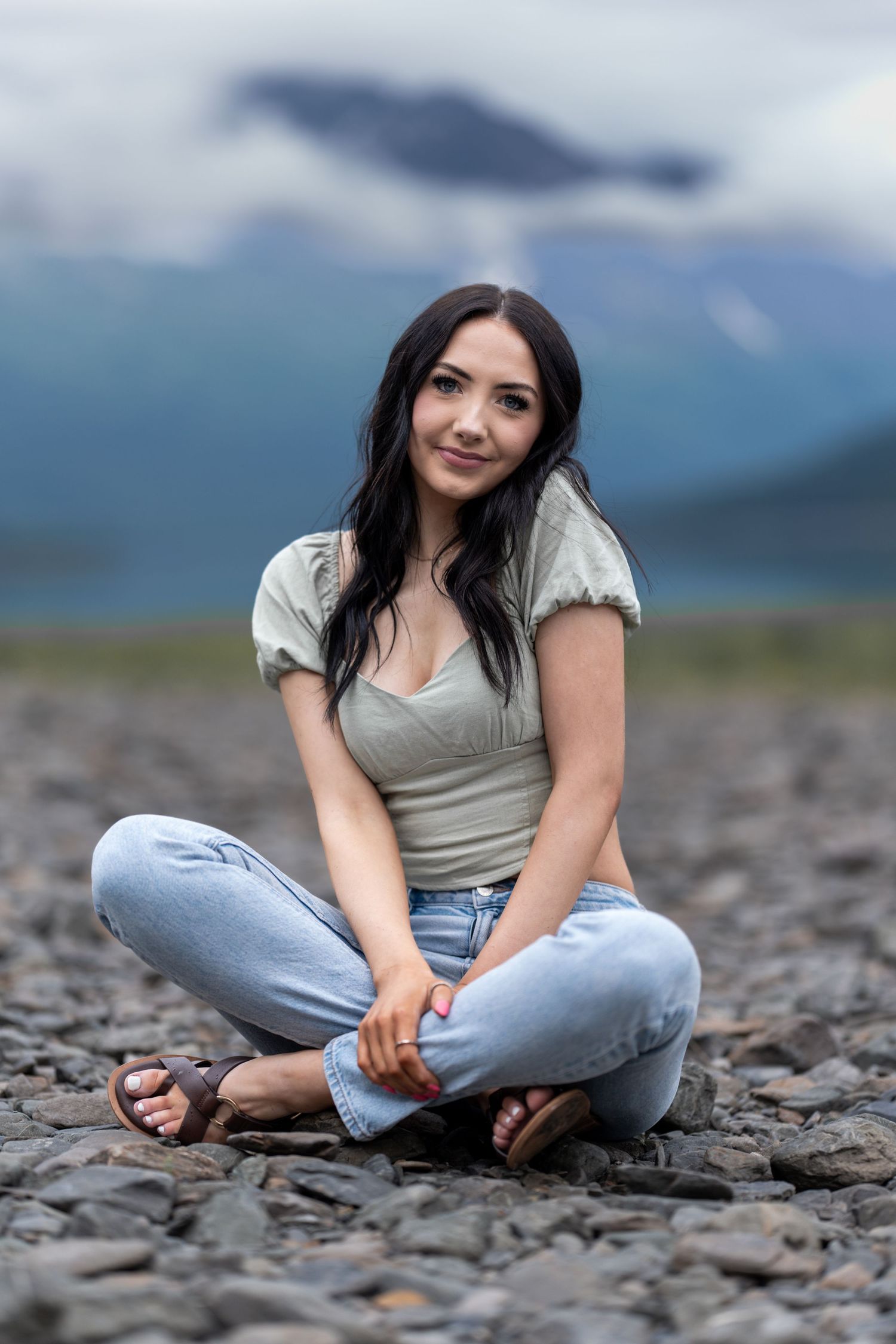 Young woman with dark hair sits cross-legged on rocky shore with misty mountains in background.