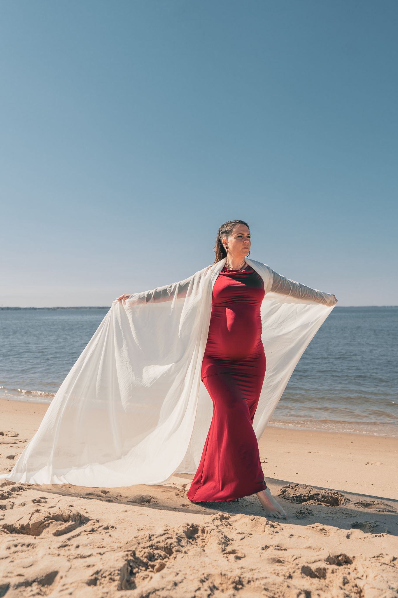 Pregnant woman in red gown holds white fabric on sunny beach by the ocean.