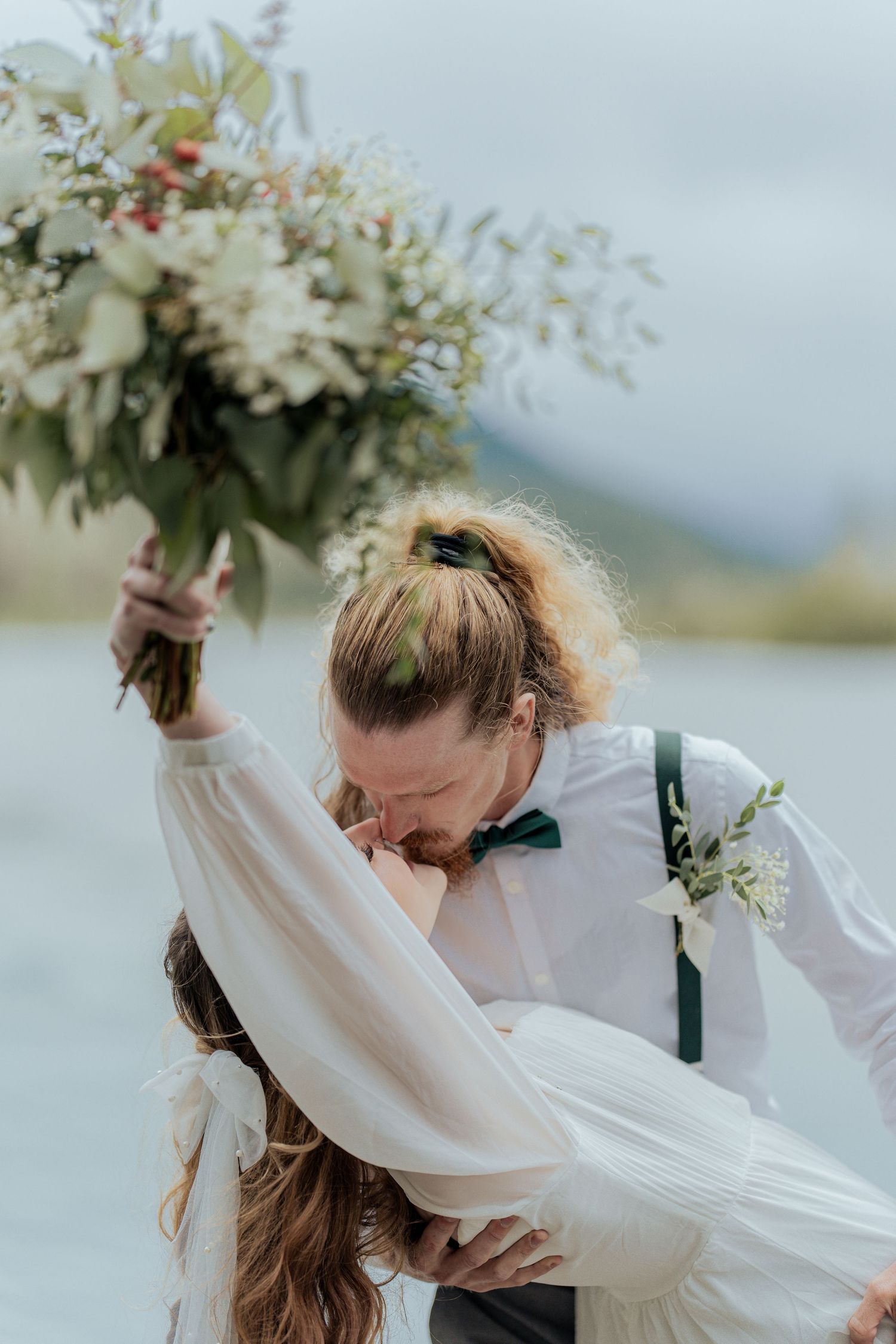 Groom dips bride near lake as she raises a white and green floral bouquet overhead.