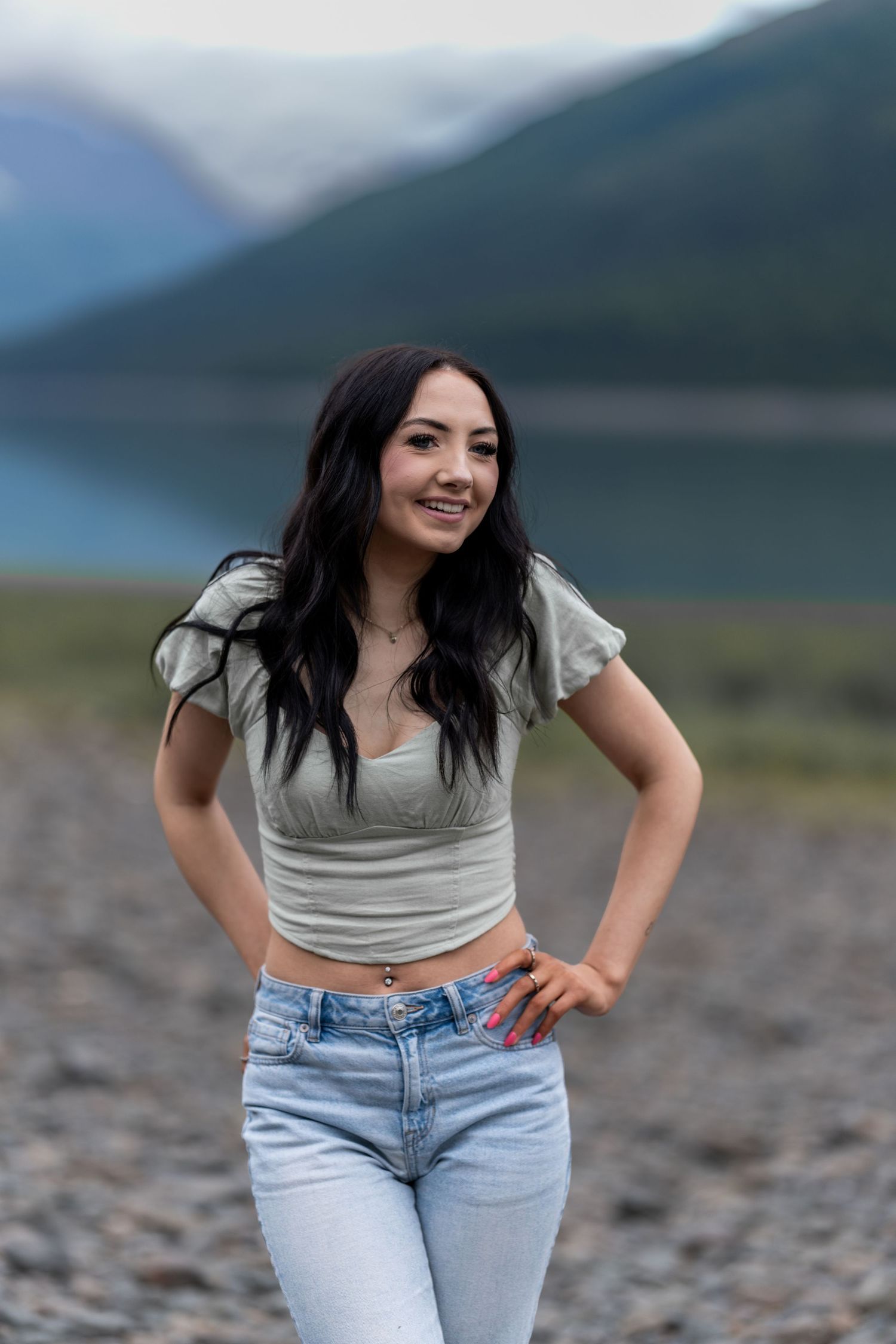 Smiling dark-haired woman in crop top and jeans poses near mountain lake.