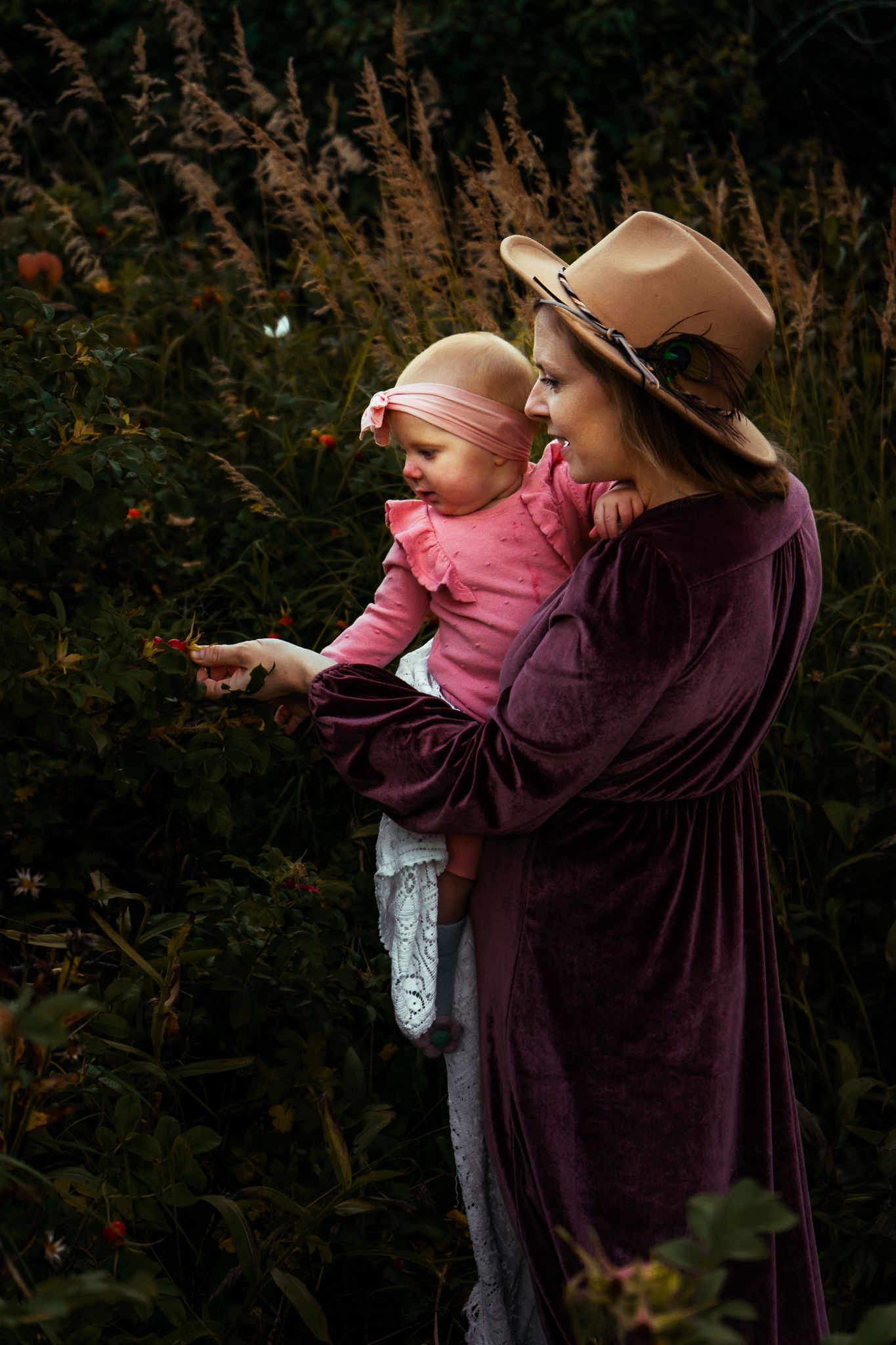 Mother in burgundy velvet dress and tan hat shows baby in pink a flower in lush garden.