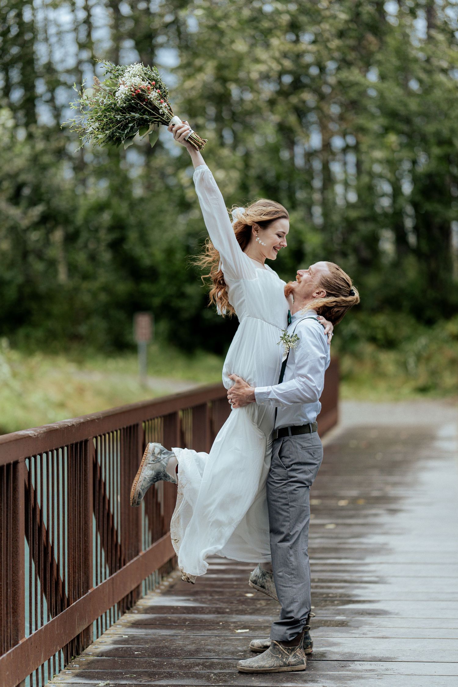 Groom lifts bride on wooden bridge as she raises bouquet joyfully with forest backdrop.