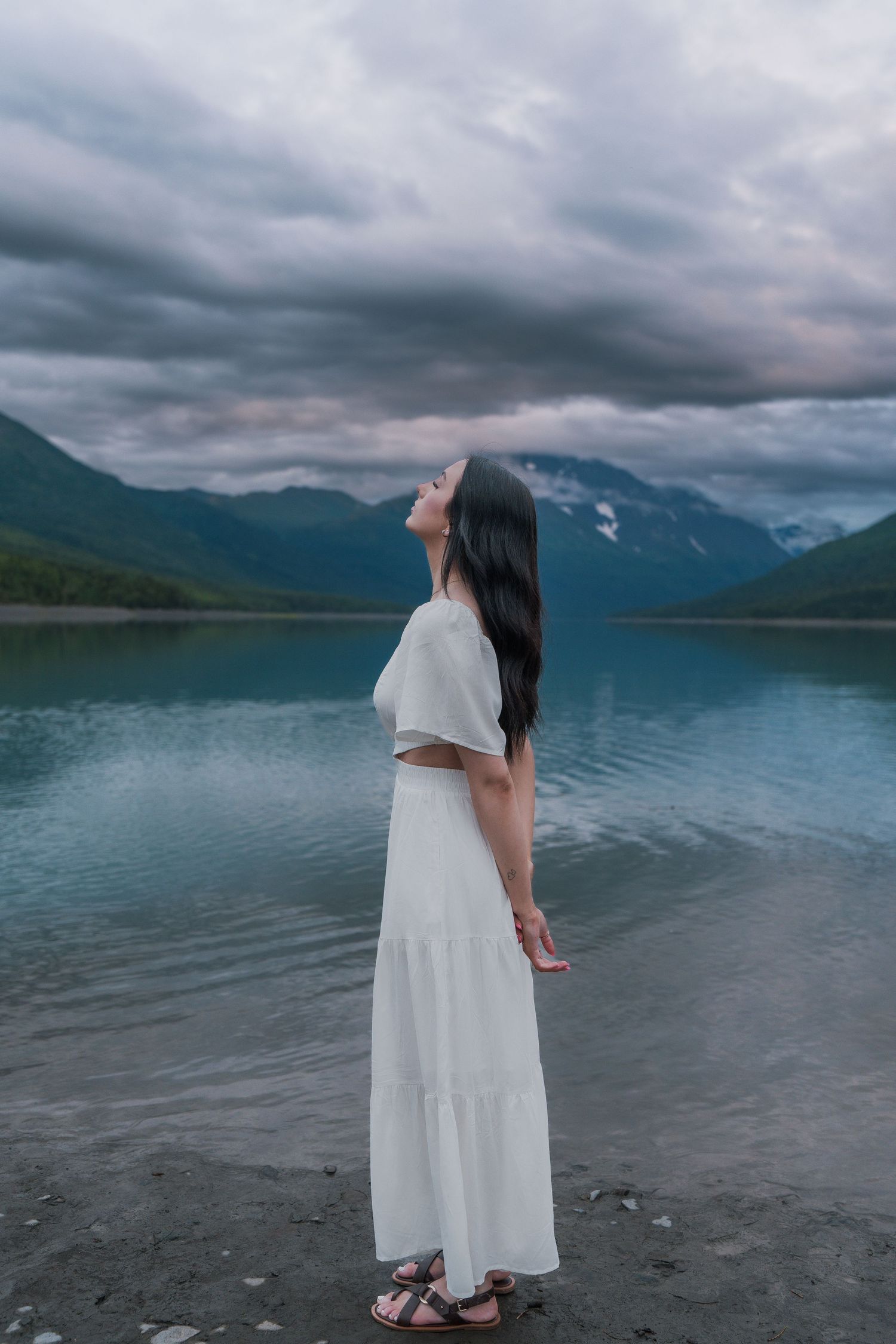 Woman in white dress looks up by calm mountain lake under cloudy evening sky.