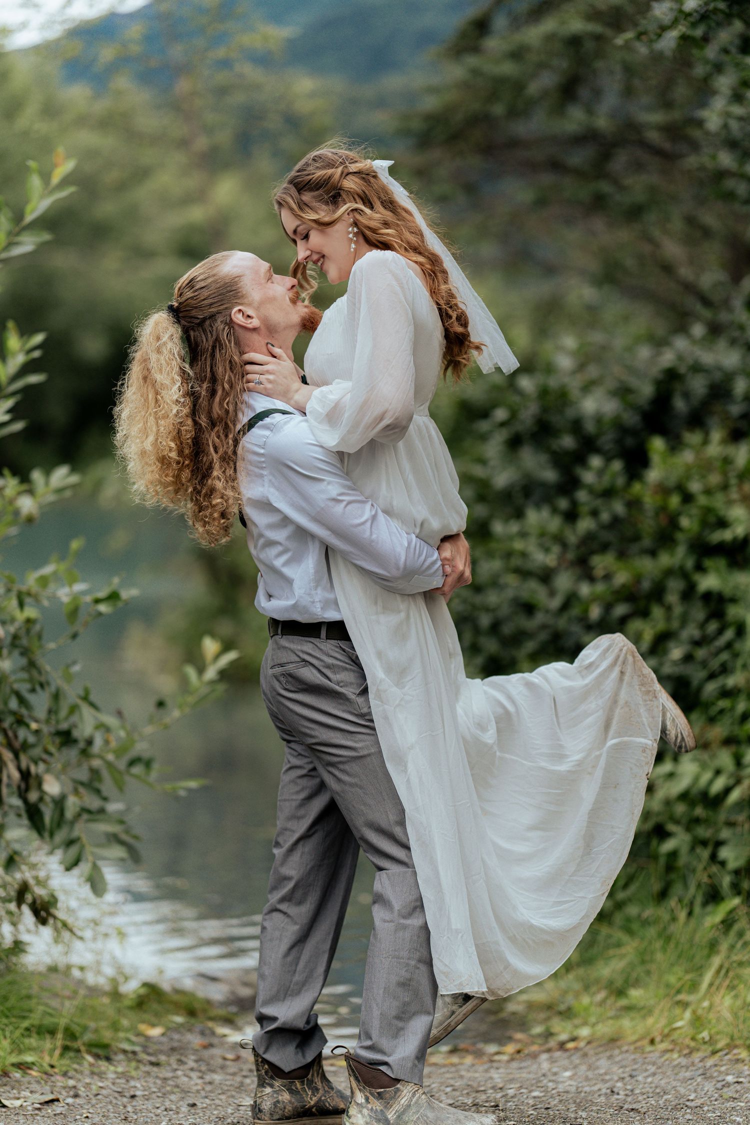 Groom lifts bride in white dress outdoors near lush greenery and a serene lake.