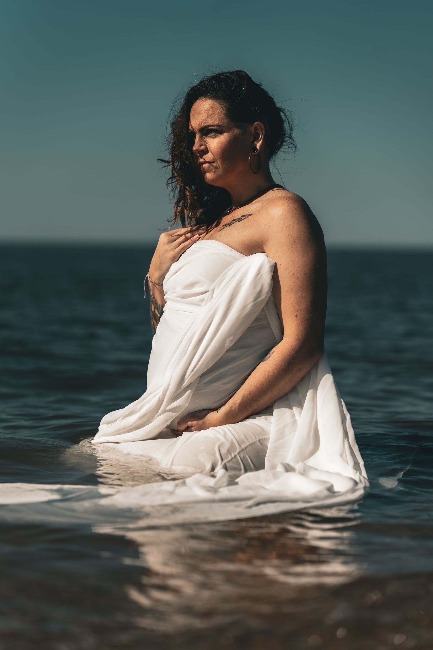 Woman wrapped in white fabric sitting in ocean water under clear blue sky in artistic portrait.