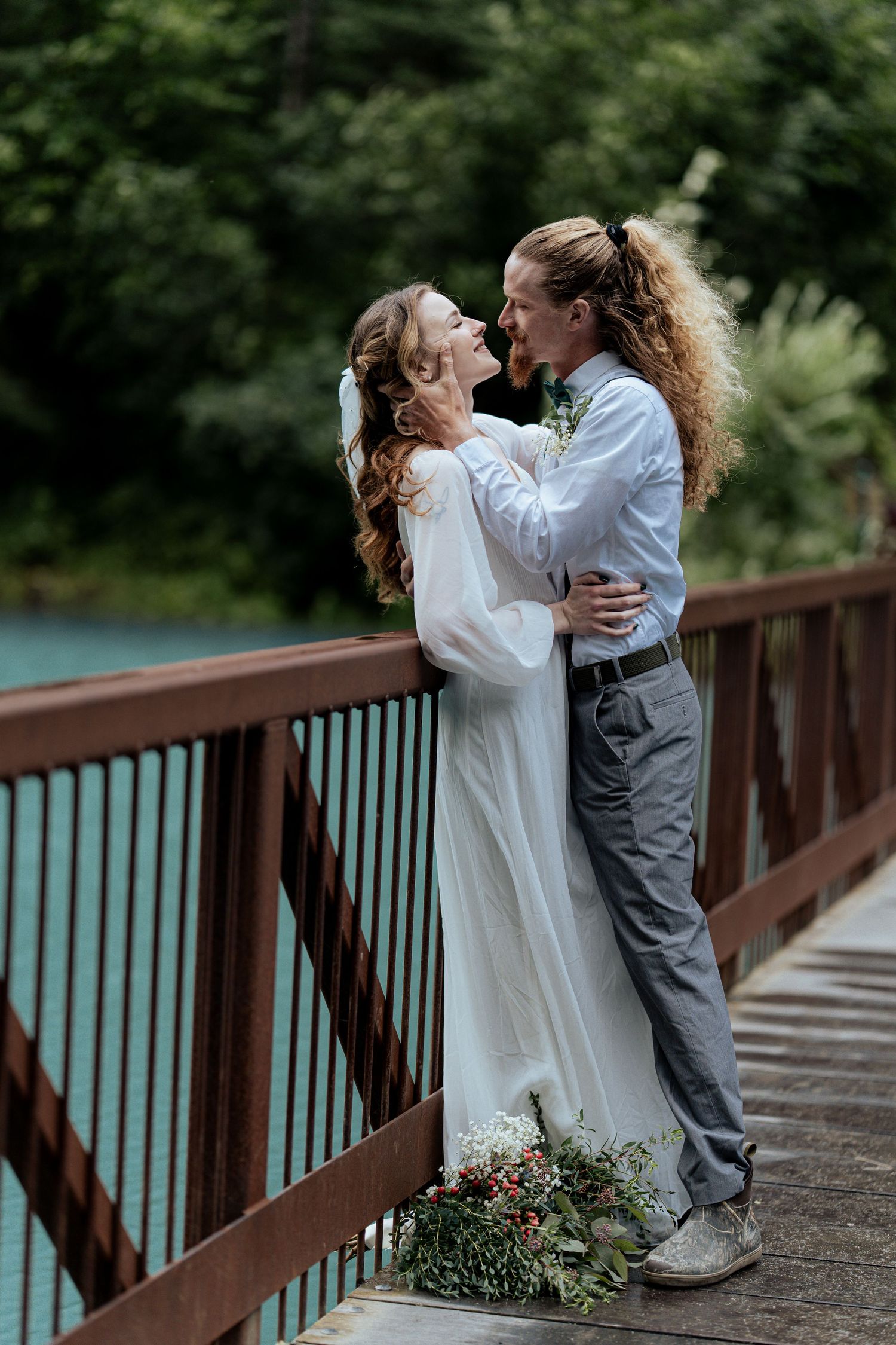 Bride and groom sharing romantic kiss on rustic bridge over turquoise river in forest setting.