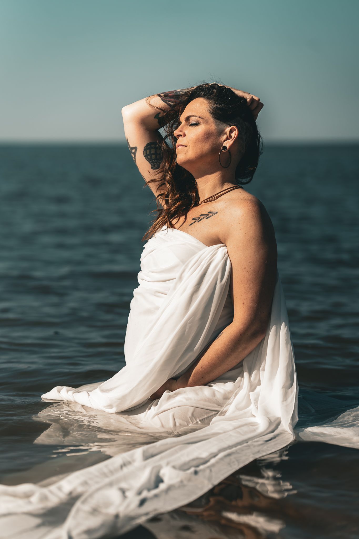 Tattooed woman wrapped in white fabric sitting in ocean water with eyes closed in serene pose.