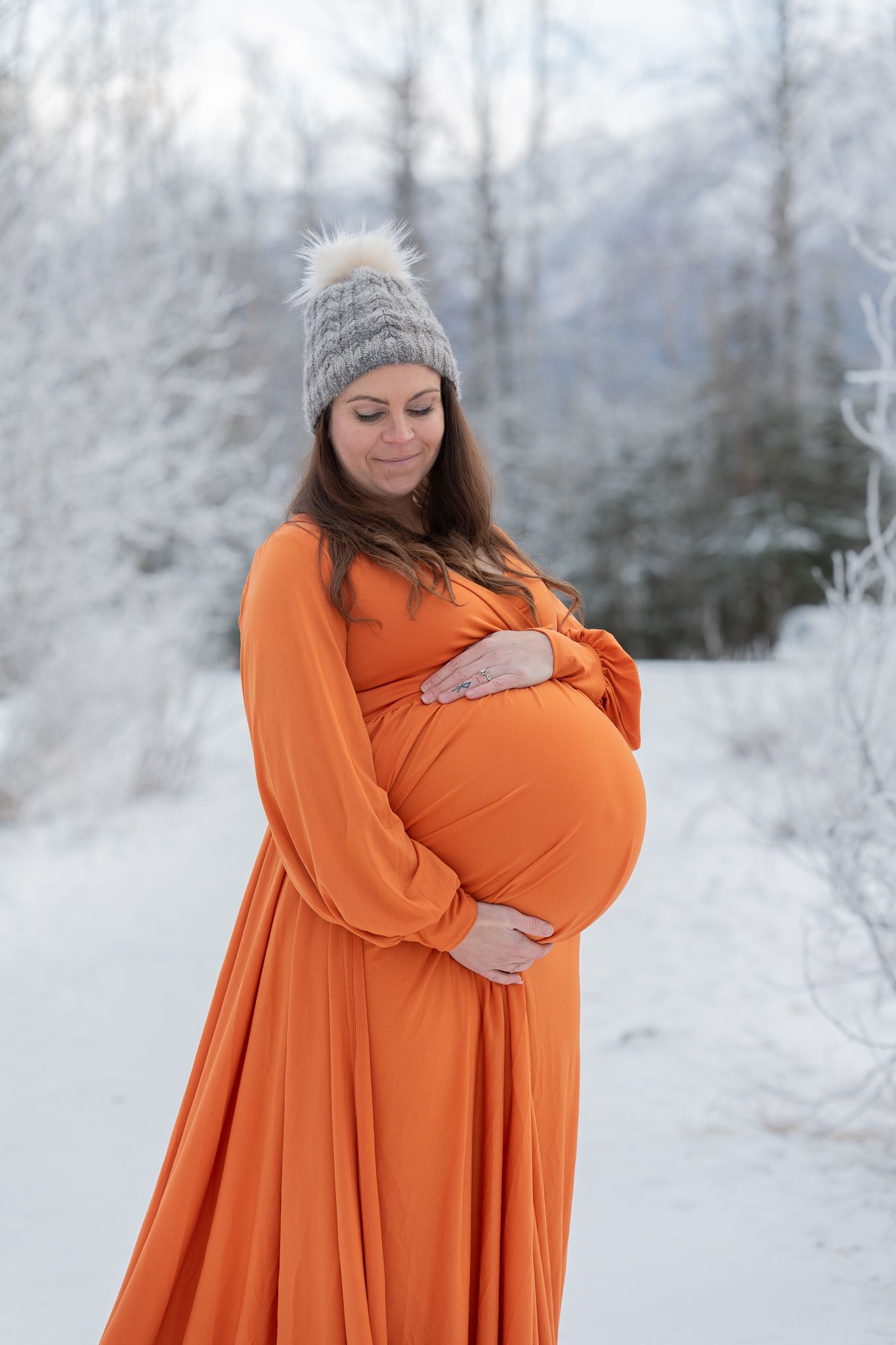 A smiling pregnant woman in an orange gown and knit hat cradles her bump in a snowy winter forest.