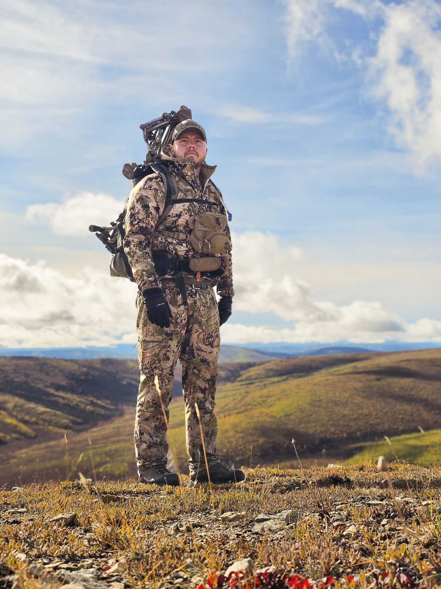 Hunter in full camouflage gear standing on mountain hilltop with vast wilderness landscape behind him.