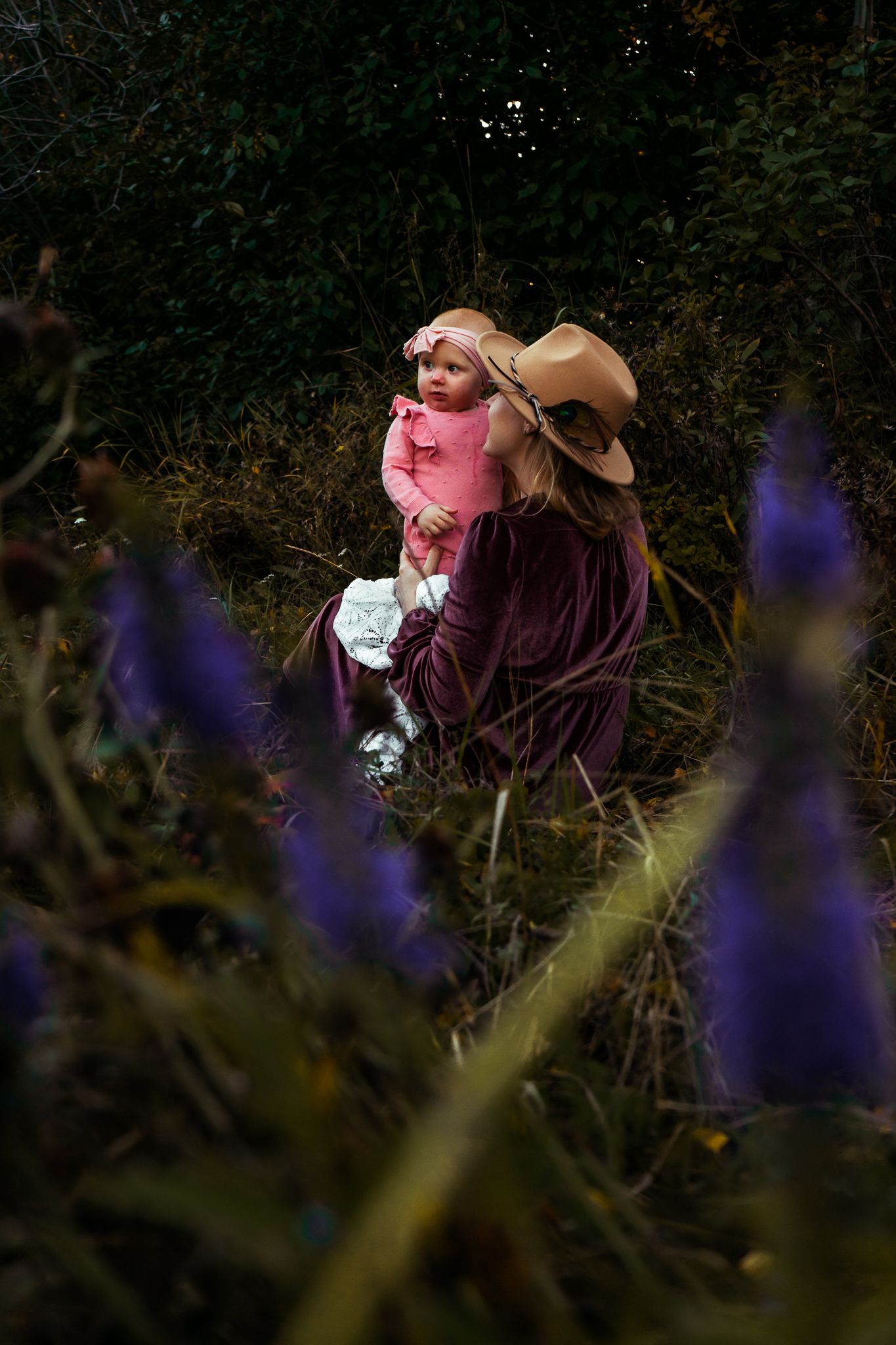 Woman in purple velvet dress and tan hat holds baby in pink among wildflowers and dark foliage.