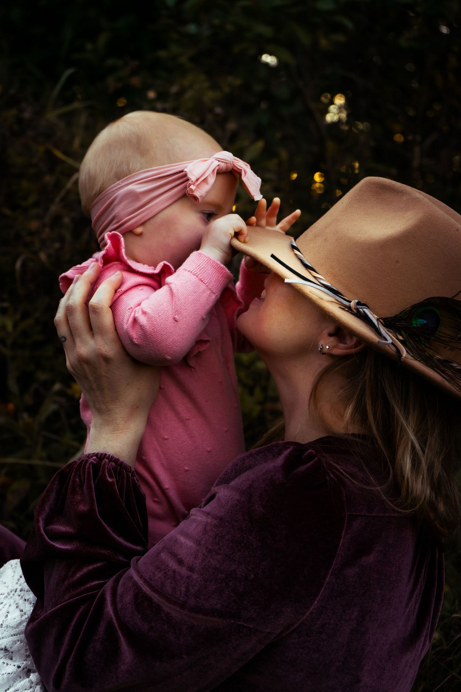 Mother in wide-brim hat tenderly kissing her baby dressed in pink against green foliage background.