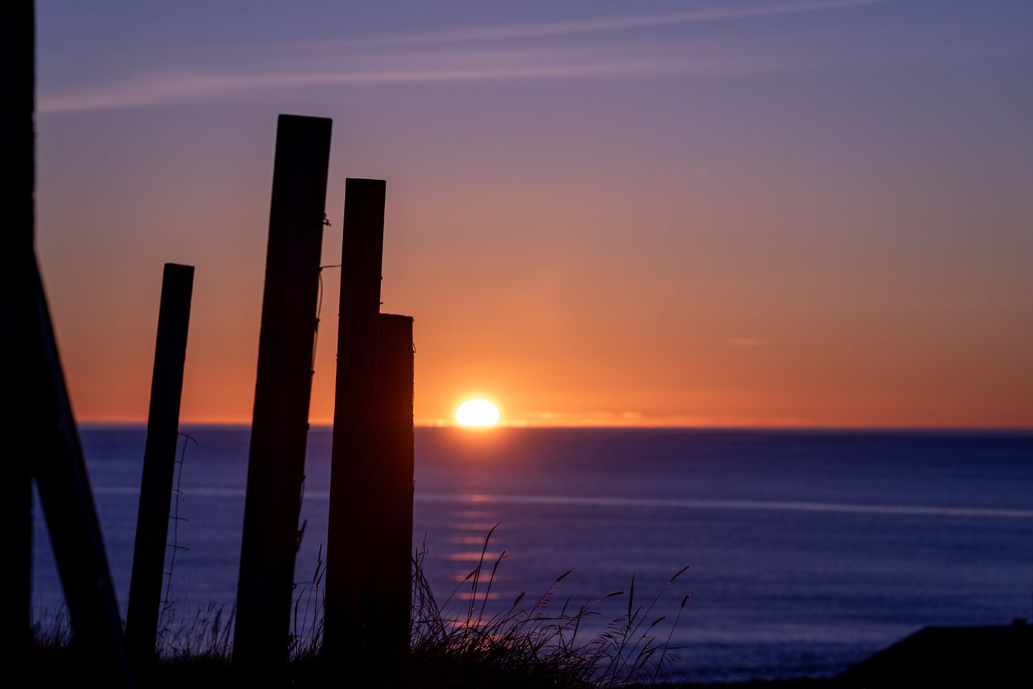 Silhouetted wooden fence posts against vibrant purple and orange ocean sunset sky.