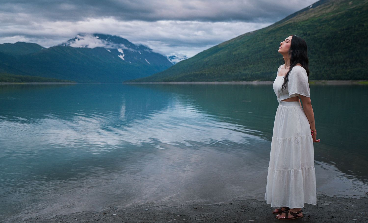 Woman in white dress standing by serene mountain lake with snow-capped peaks reflected in water.