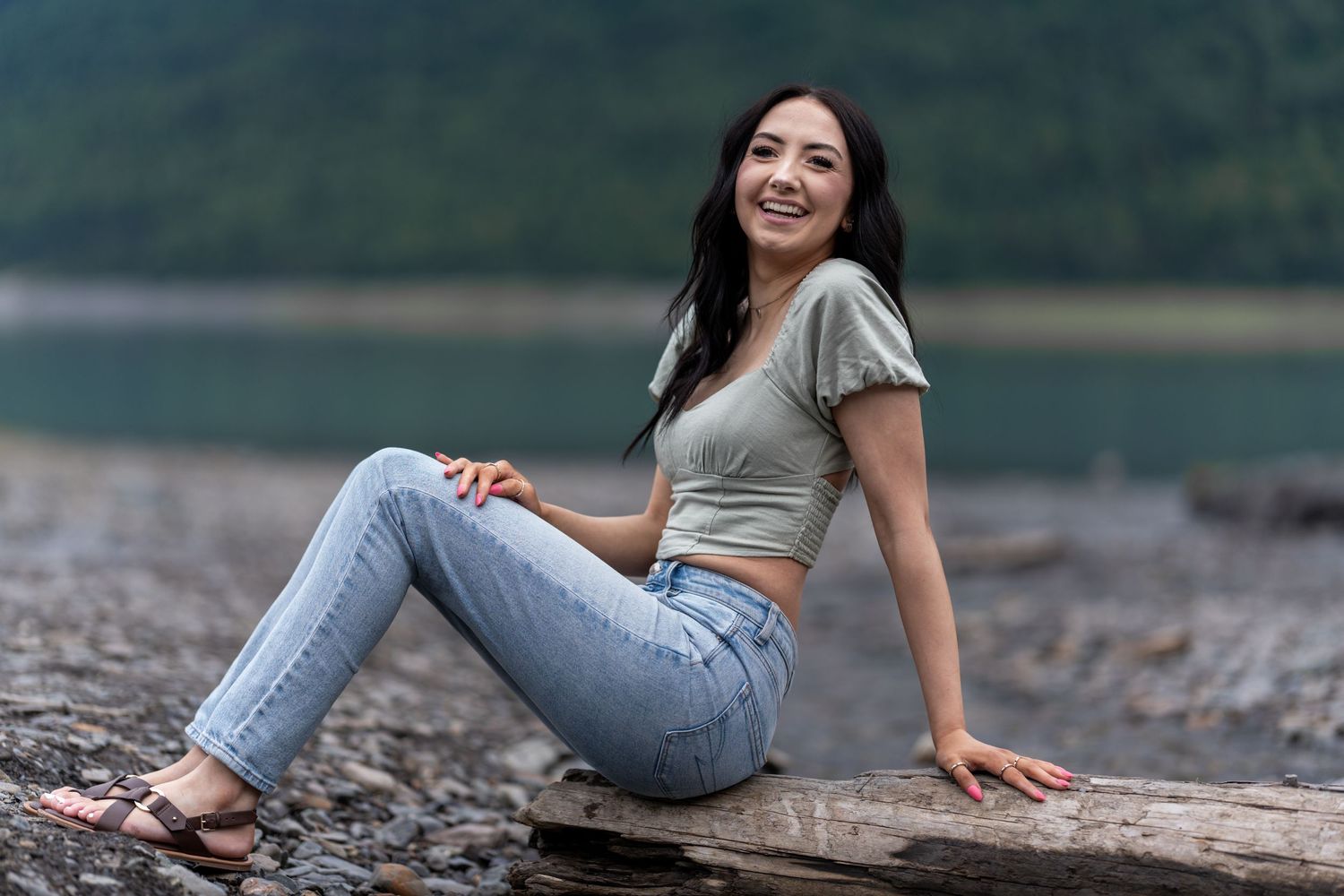 Laughing brunette woman in jeans and crop top sits on driftwood by a scenic mountain lake.