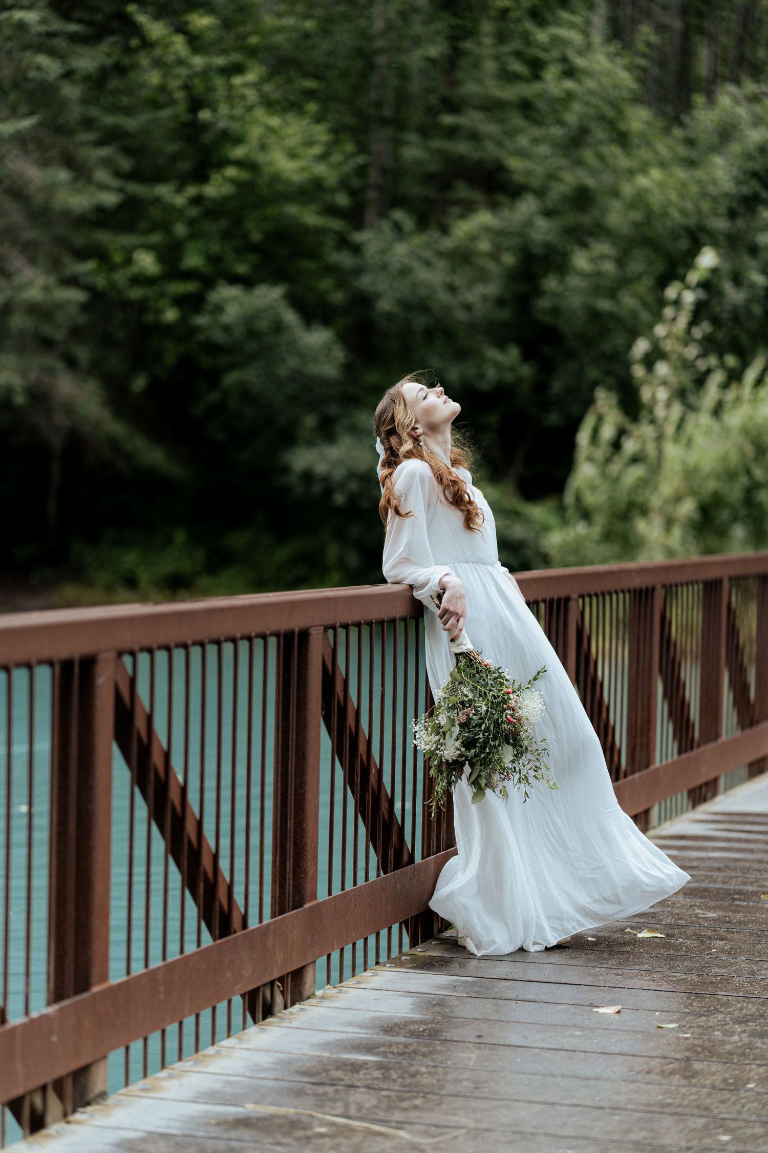Bride in white gown leans on red bridge railing gazing upward beside a turquoise river.