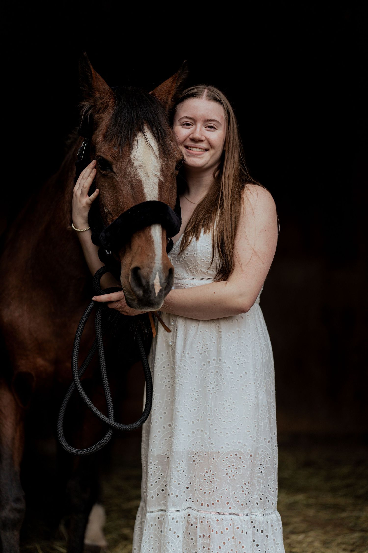 Smiling young woman in white lace dress posing with brown horse in dark stable setting.