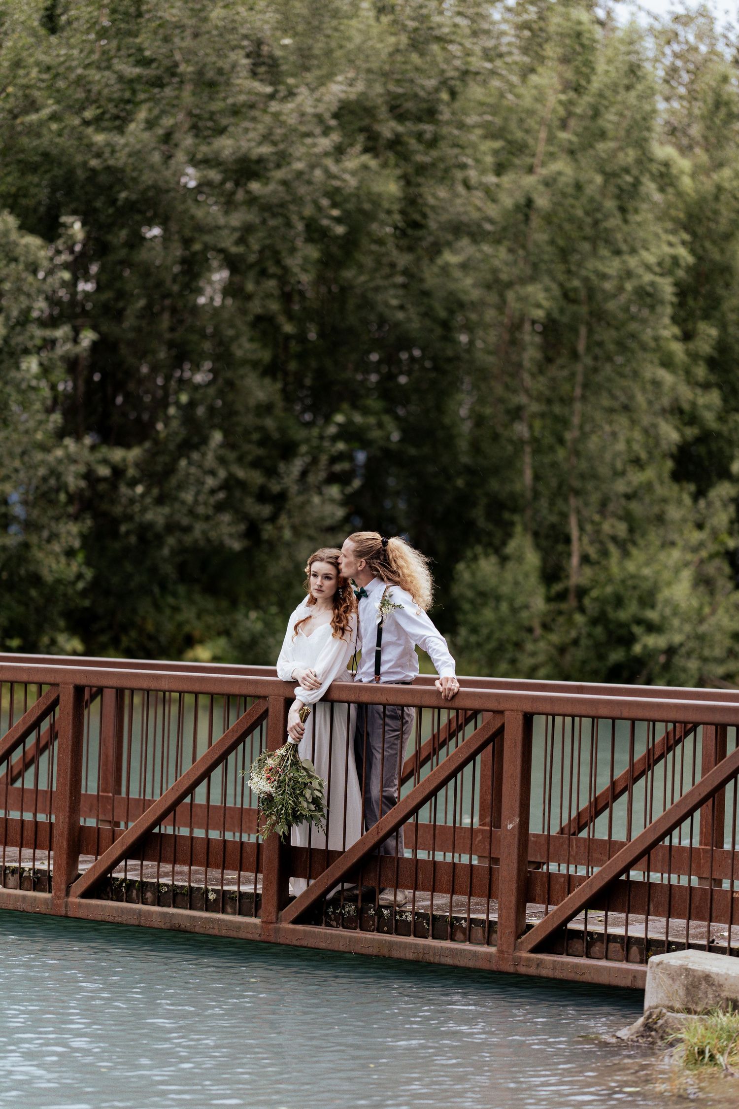 Wedding couple embracing on a red bridge over turquoise water surrounded by green trees.
