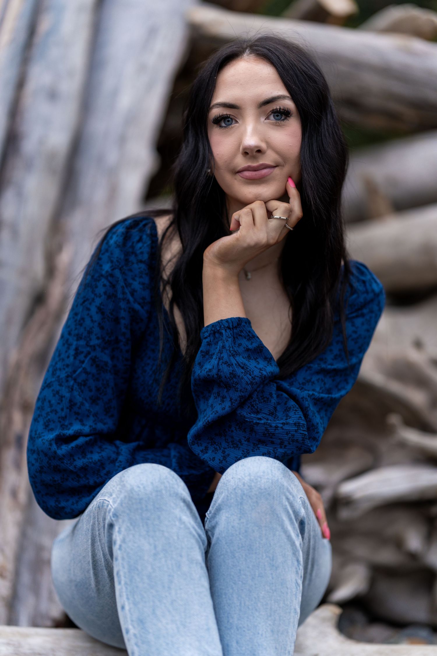 Dark-haired woman in blue top and jeans sits thoughtfully on driftwood logs.