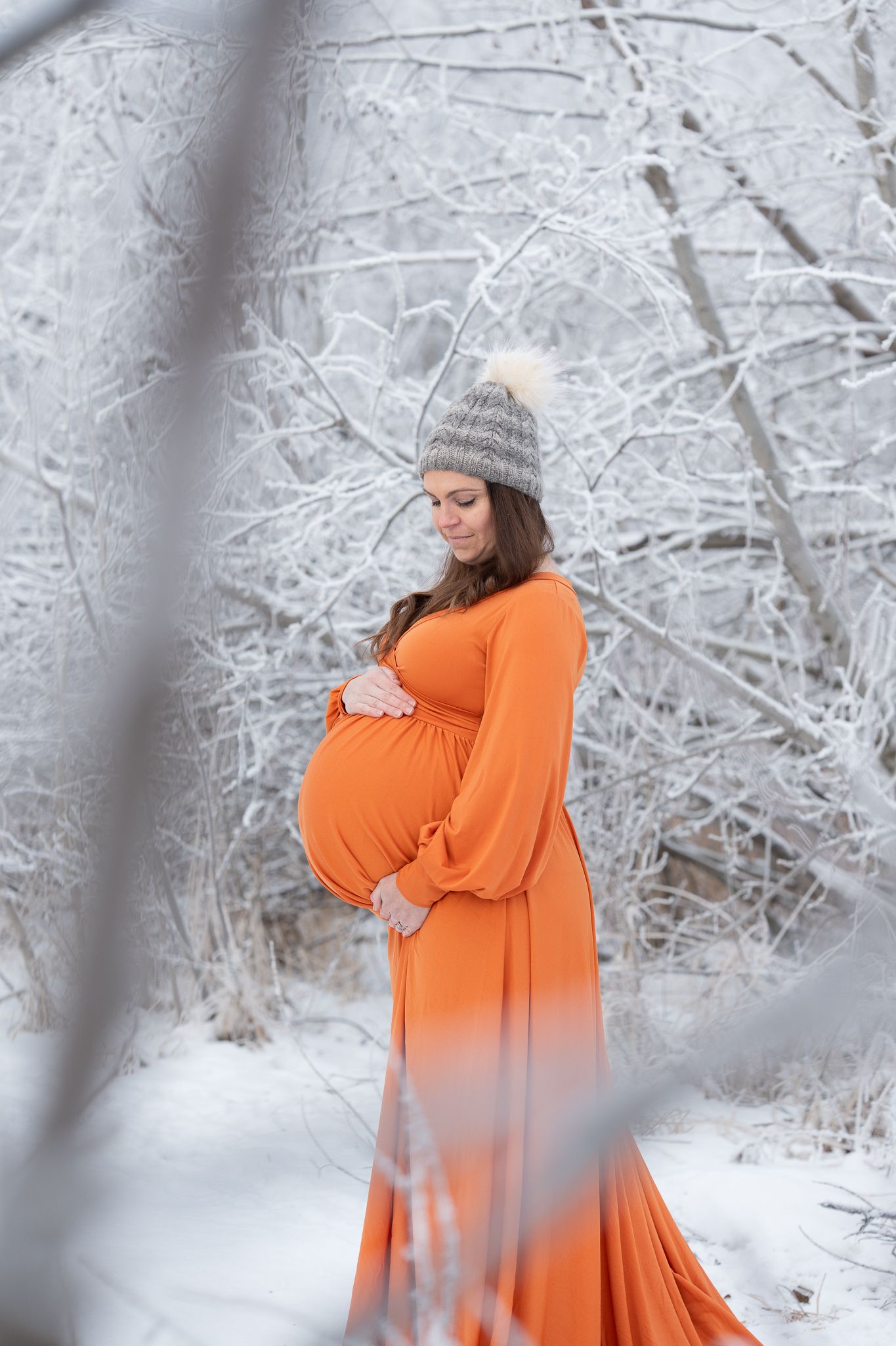 Pregnant woman in orange dress cradles belly amid frost-covered trees in winter.