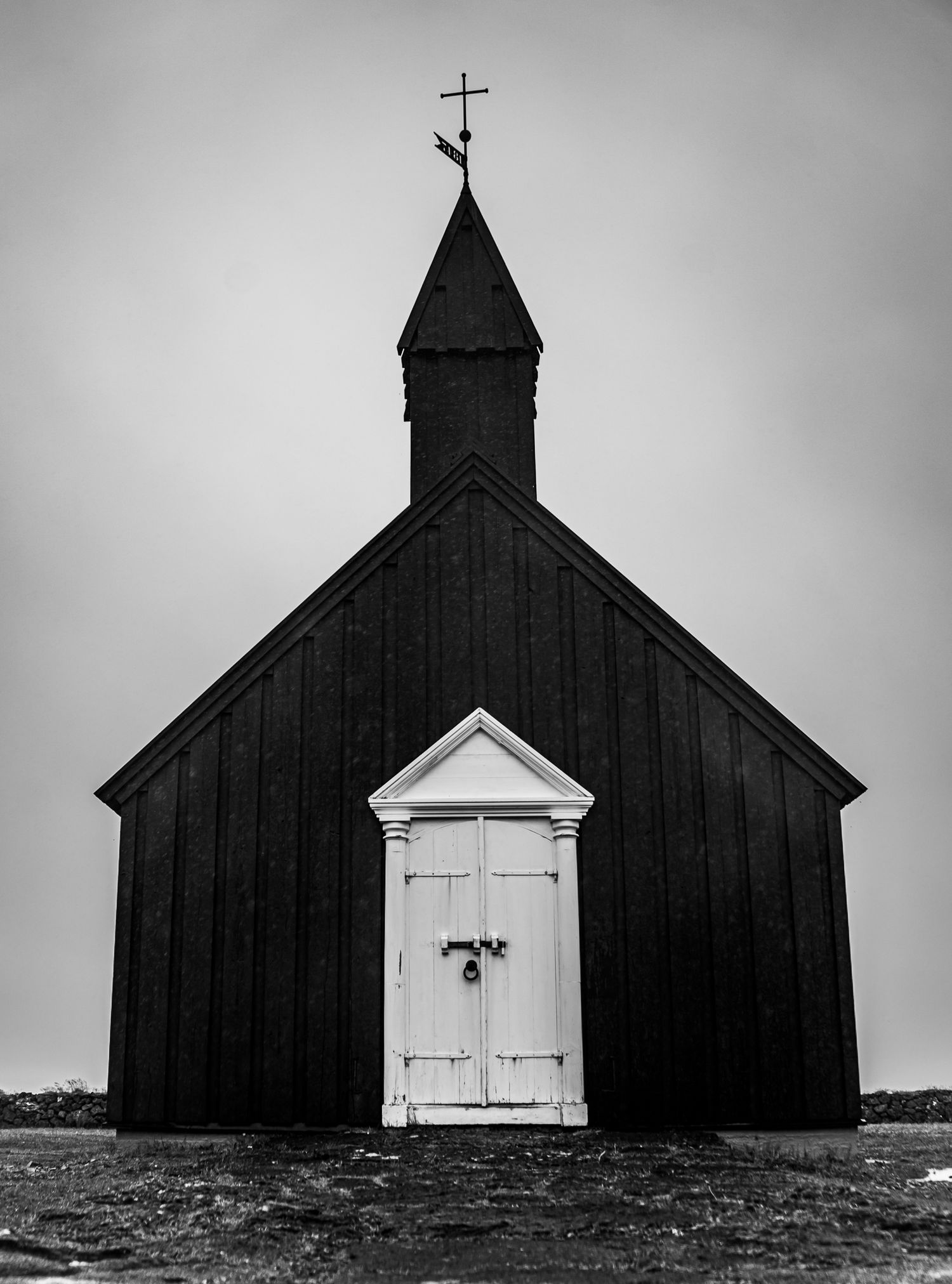 Black wooden church with white door and cross steeple in dramatic black and white photography.