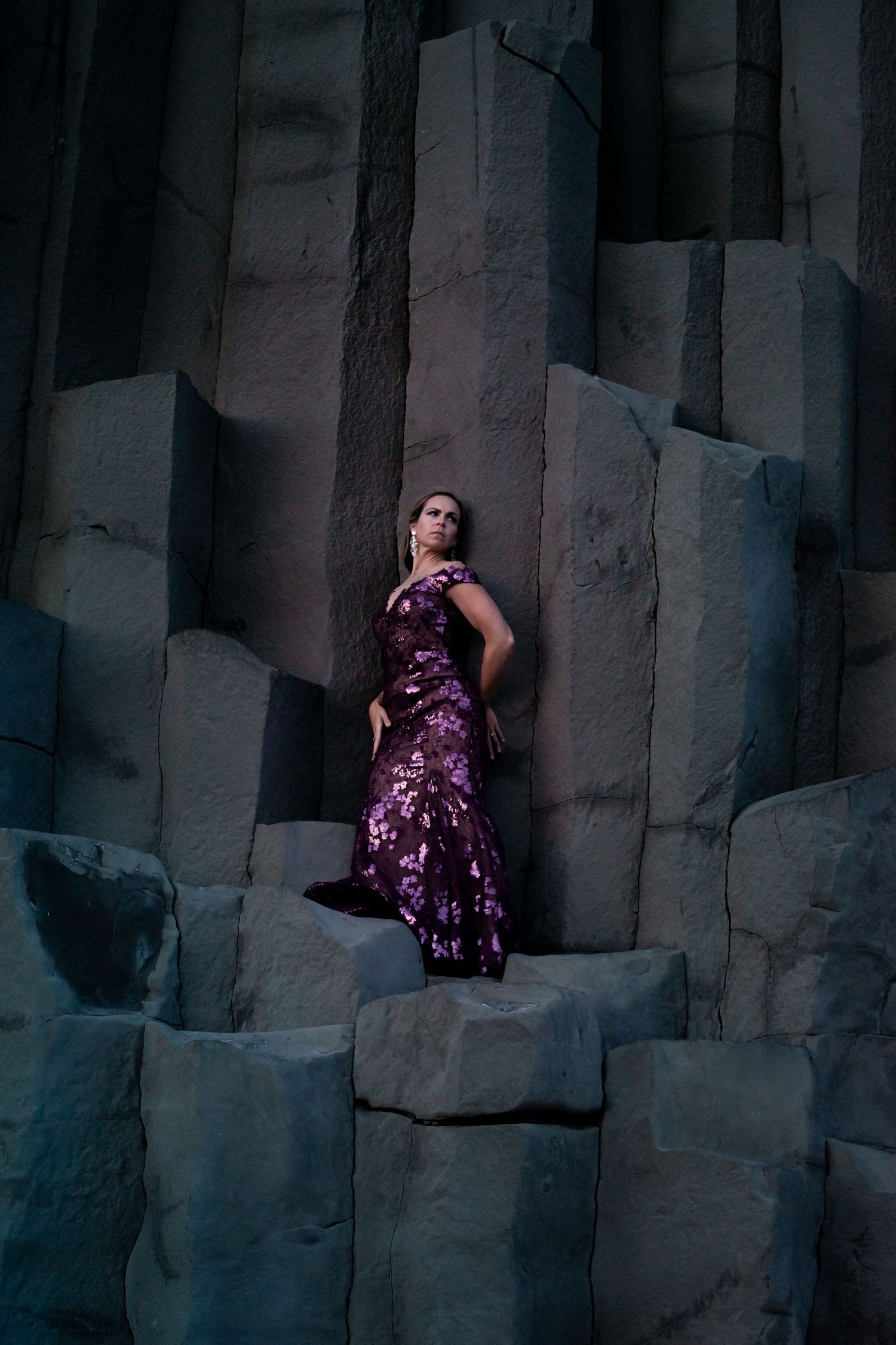 Woman in purple sequin gown posing dramatically against dark basalt column rock formations.