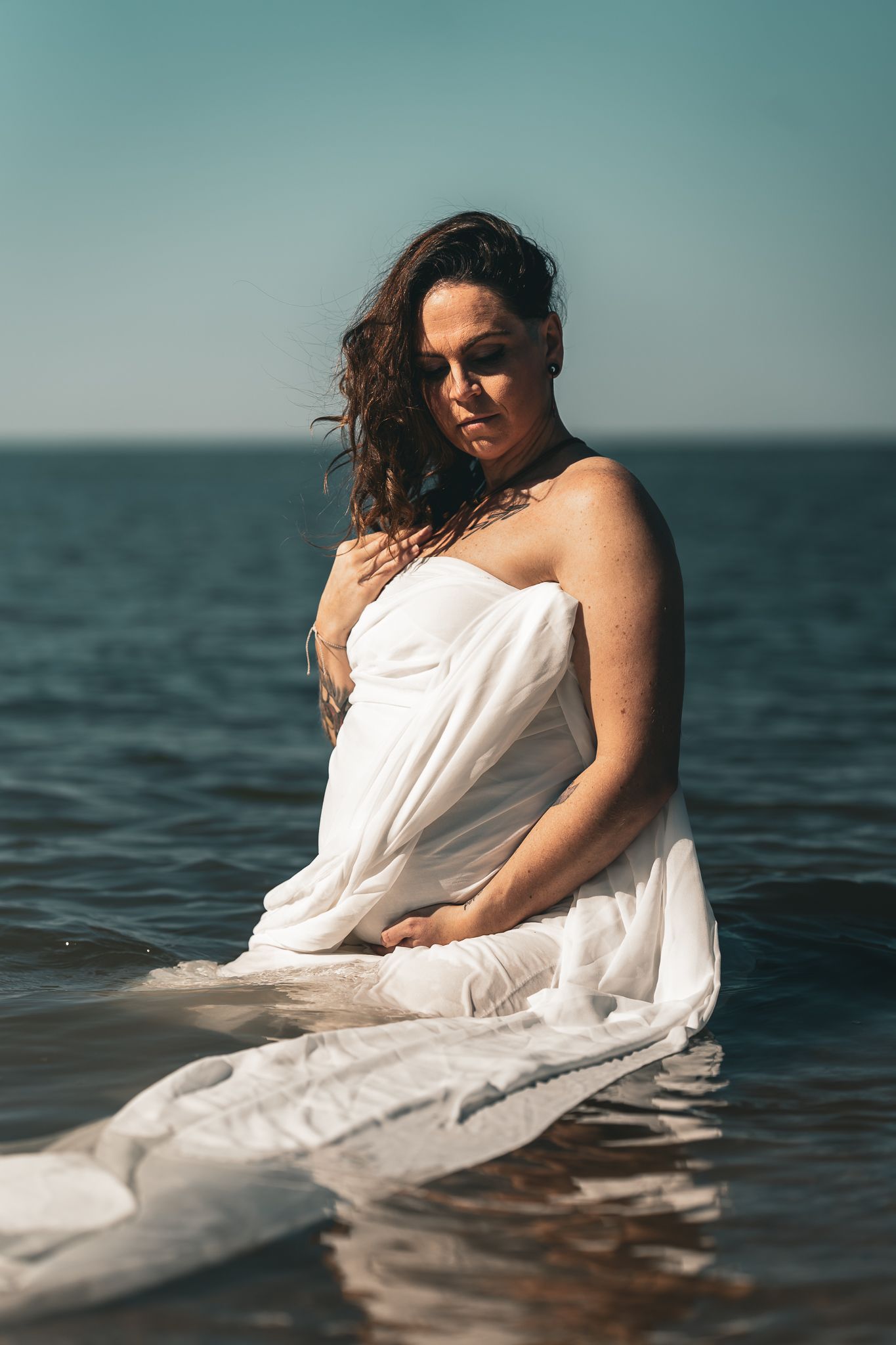 A woman wrapped in a flowing white fabric stands in ocean water during a maternity photo shoot.