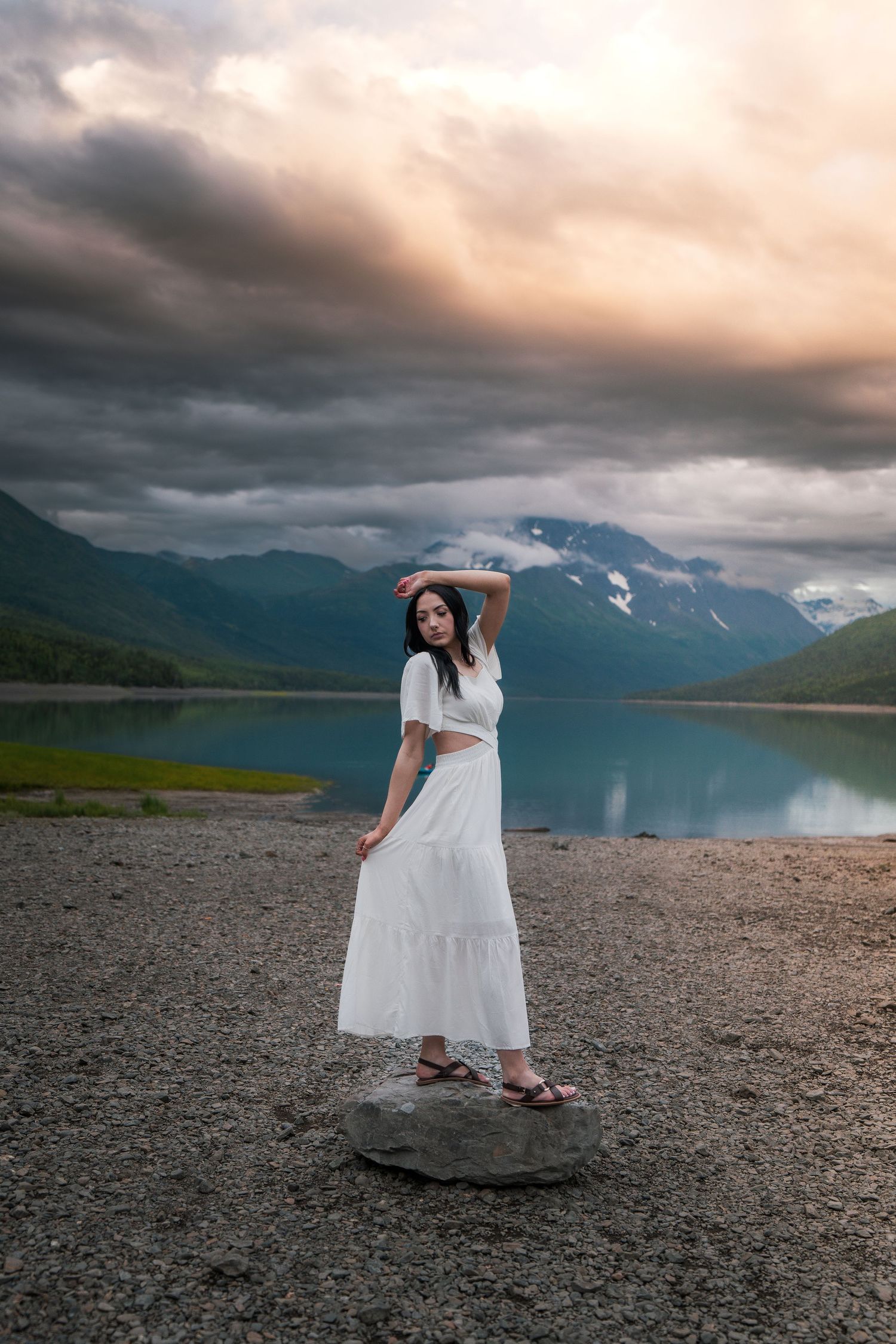 Woman in white crop top skirt stands on rock by turquoise mountain lake at dusk.