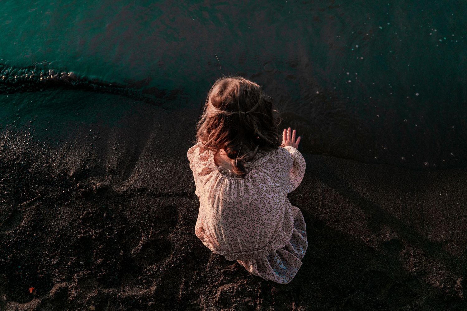 Young girl in white dress sitting on dark sand beach beside glowing turquoise water, aerial view.