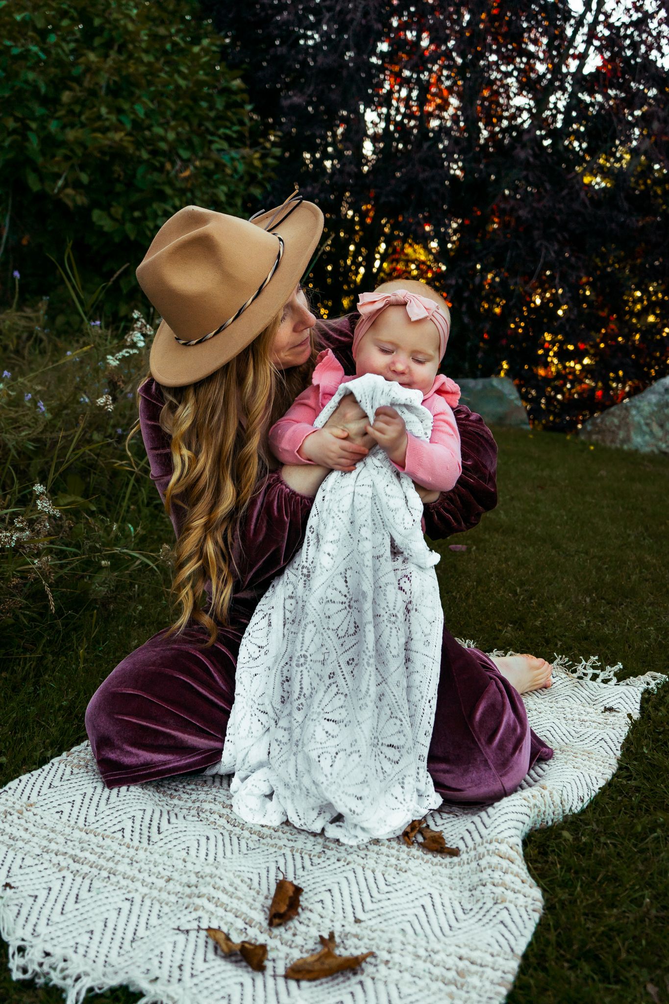 Mother in burgundy velvet dress and tan hat sits on blanket outdoors holding baby in pink outfit.