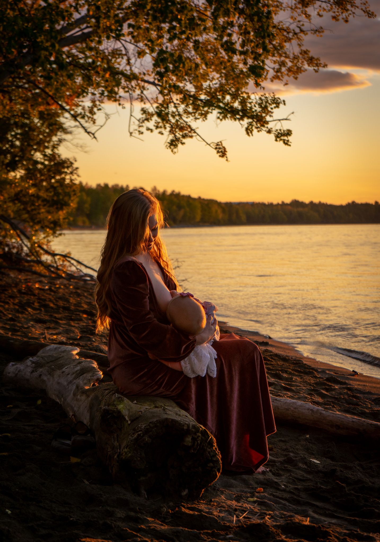 Woman in burgundy velvet dress sits on log holding baby at golden sunset by a lake.