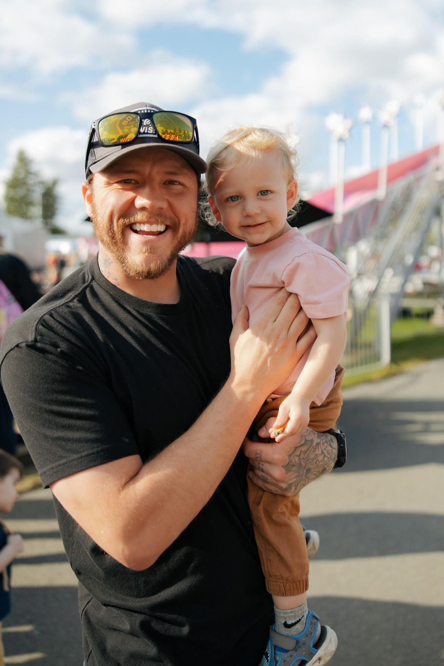 Smiling man in black cap holding a happy toddler girl at an outdoor summer event.