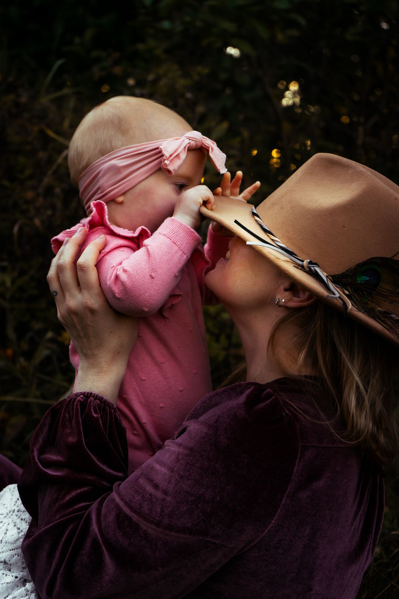 Mother in tan hat and burgundy dress lovingly kisses baby in pink outfit outdoors in nature.