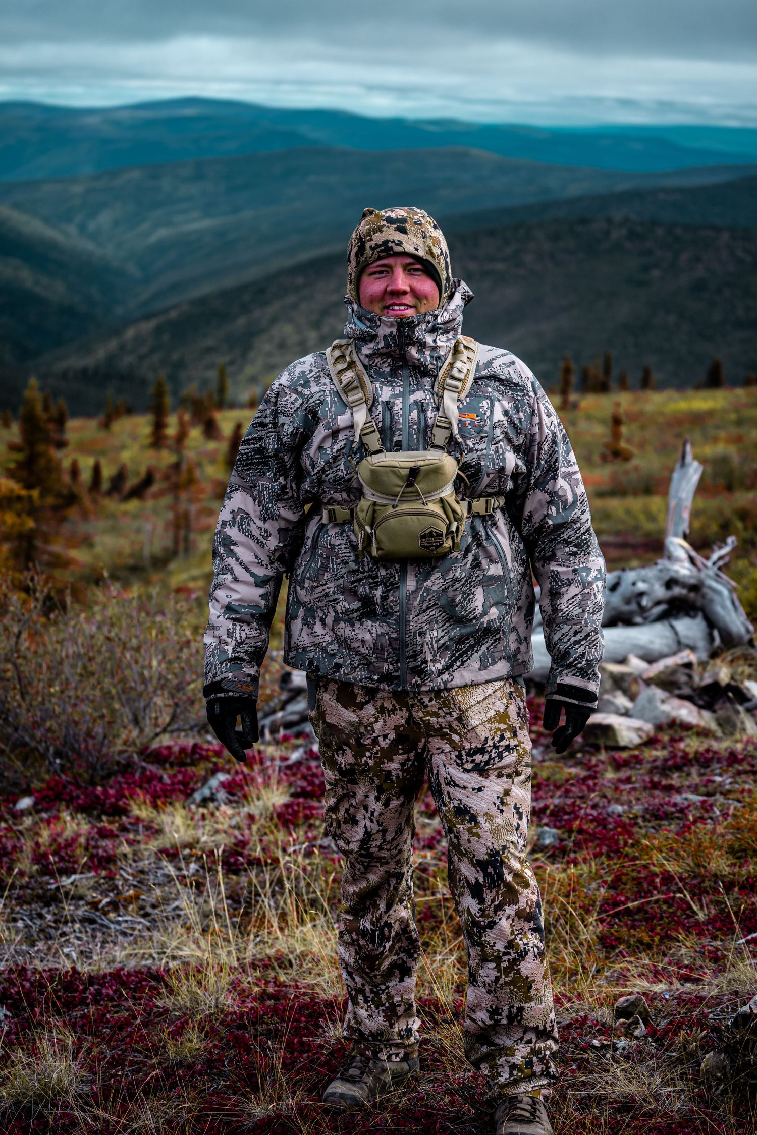 Hunter in camouflage gear smiling on Arctic tundra with vast mountain wilderness backdrop.