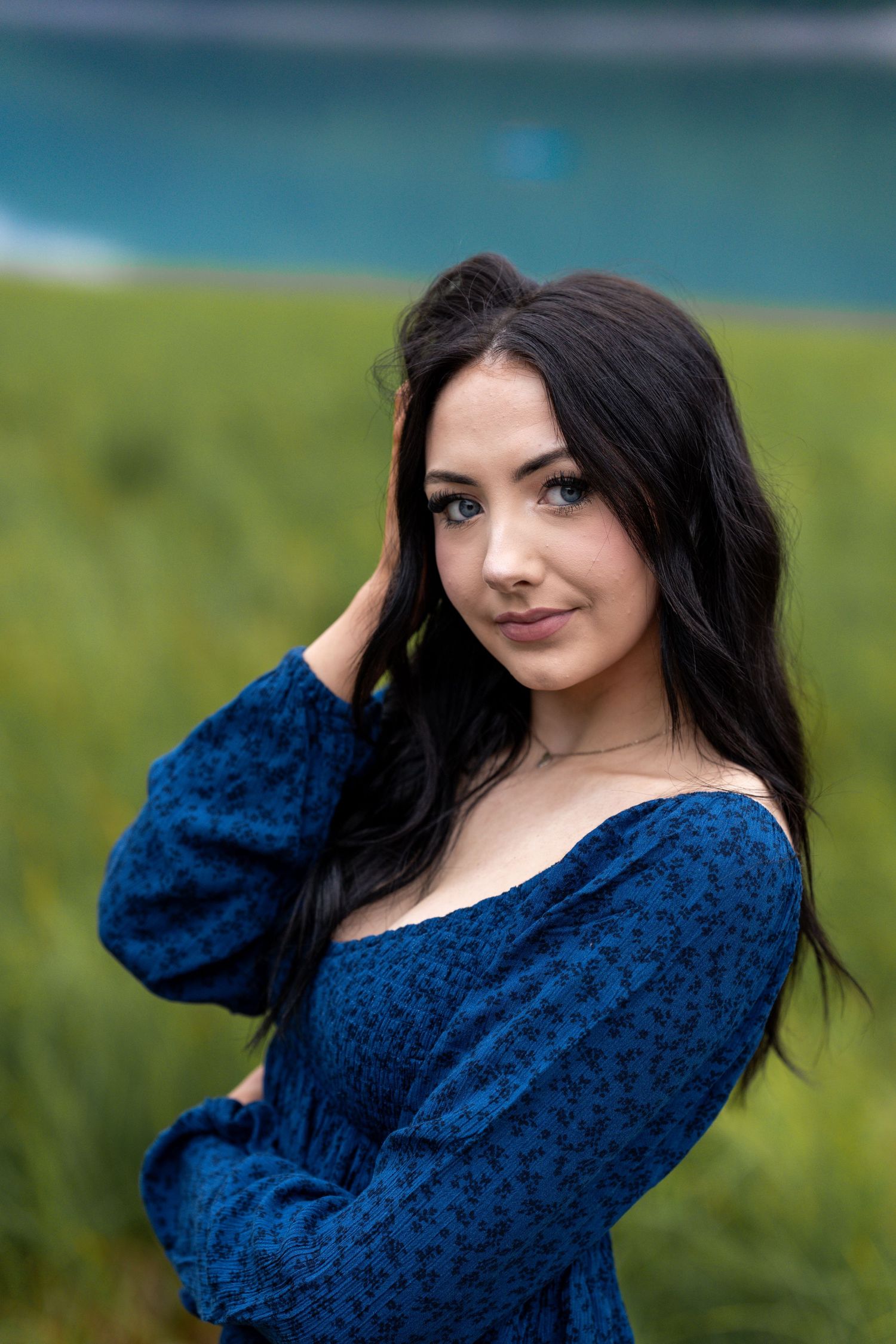 Dark-haired woman in blue floral top poses in green field near turquoise water.