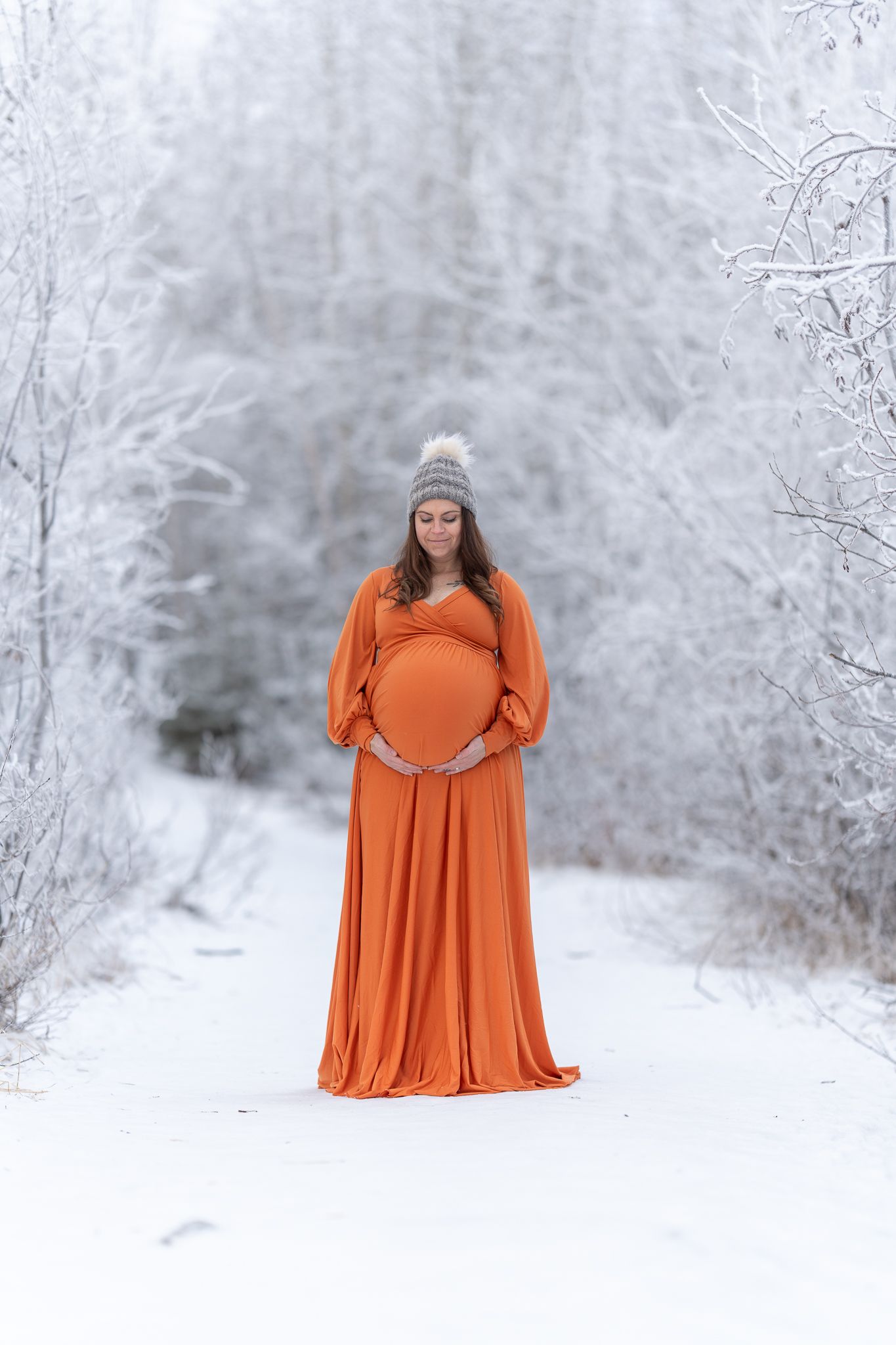 A pregnant woman in an orange gown and knit hat holds her baby bump on a snowy winter path.