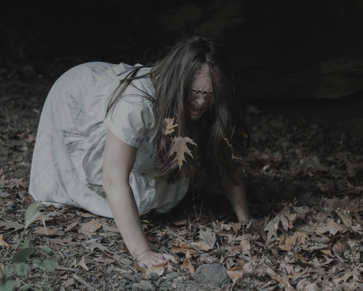 A ghostly woman in a white dress crouches on a leaf-covered forest floor with hair covering her face.