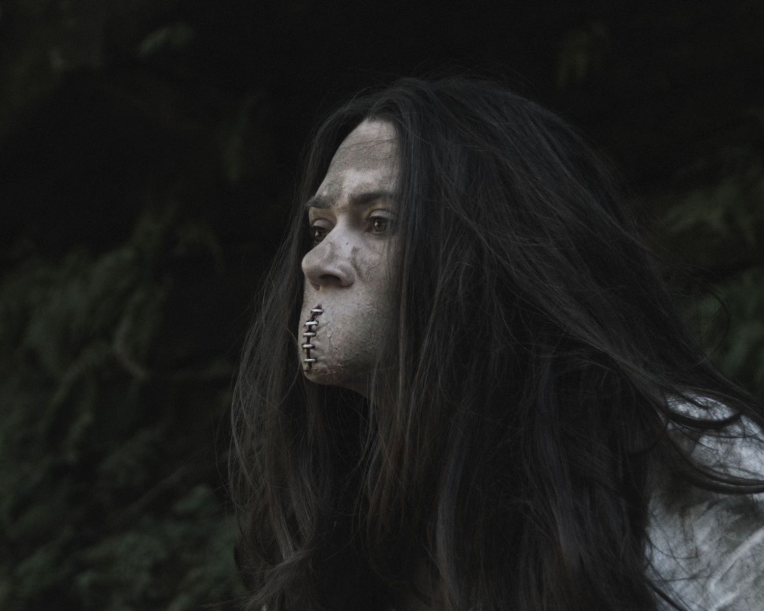 Creepy horror makeup with stitched mouth and long dark hair against a dark forest background.