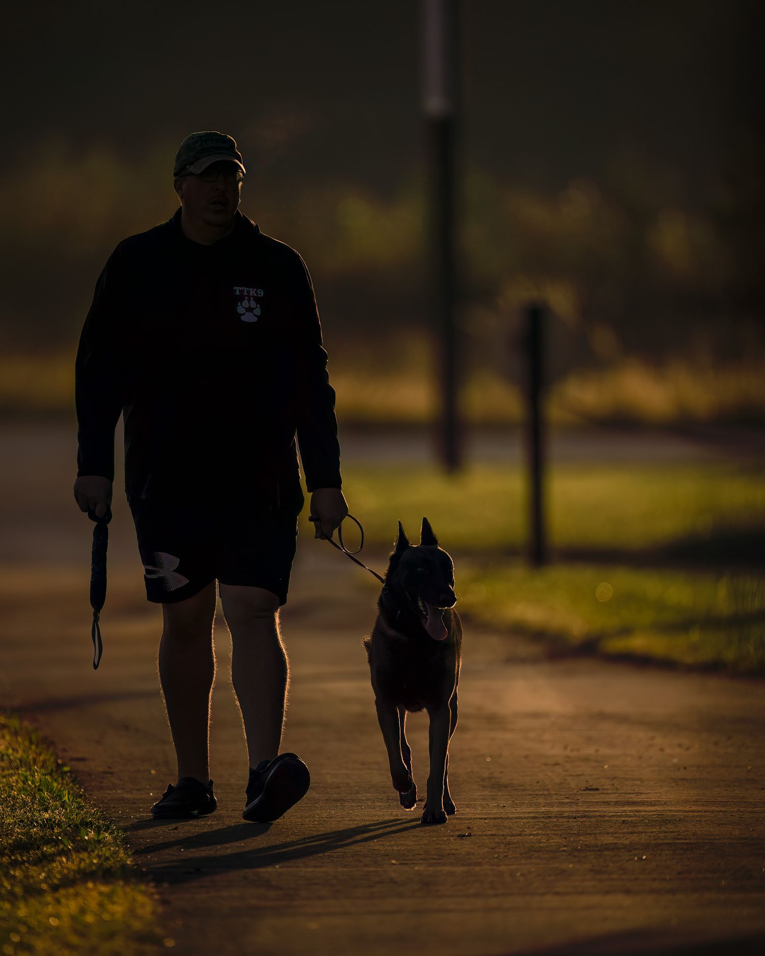 Man walking a dog on a leash along a path at dusk, silhouetted by warm golden light in a serene outdoor setting.