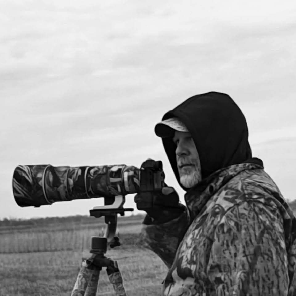 Black and white photo of a man in camouflage gear using a large telephoto lens on a tripod outdoors.
