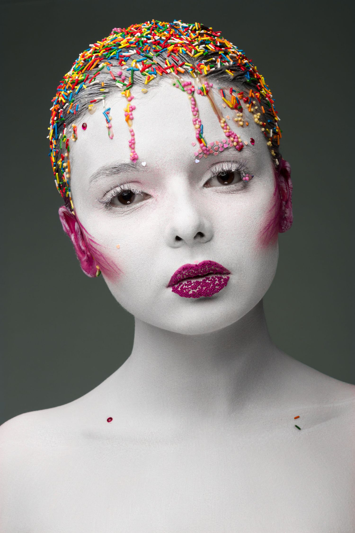 Creative portrait with white face paint, colorful sprinkles on head, and glitter pink lips against gray background.