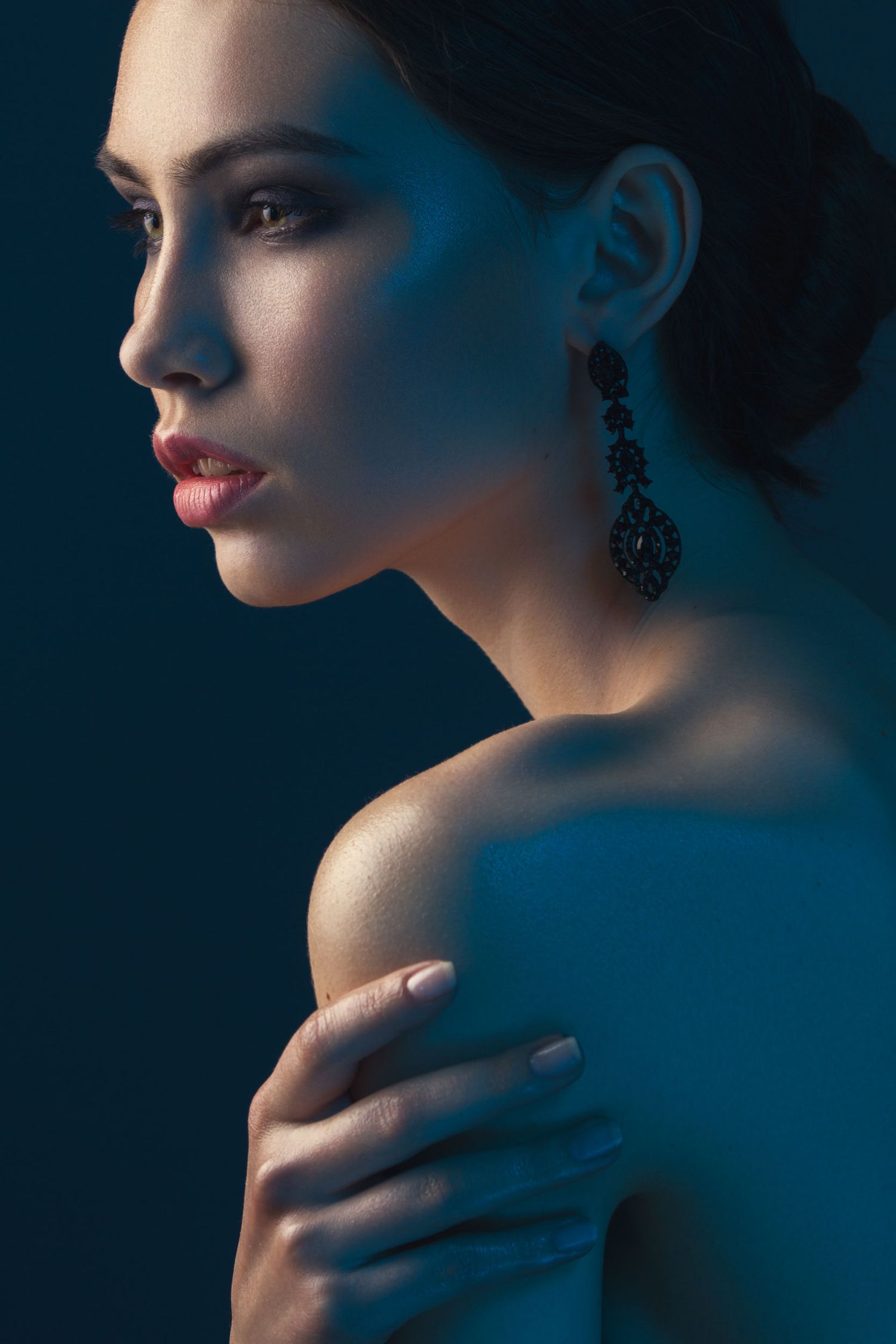 A woman in profile with elegant drop earrings, bare shoulder, and red lips against a dark blue background.