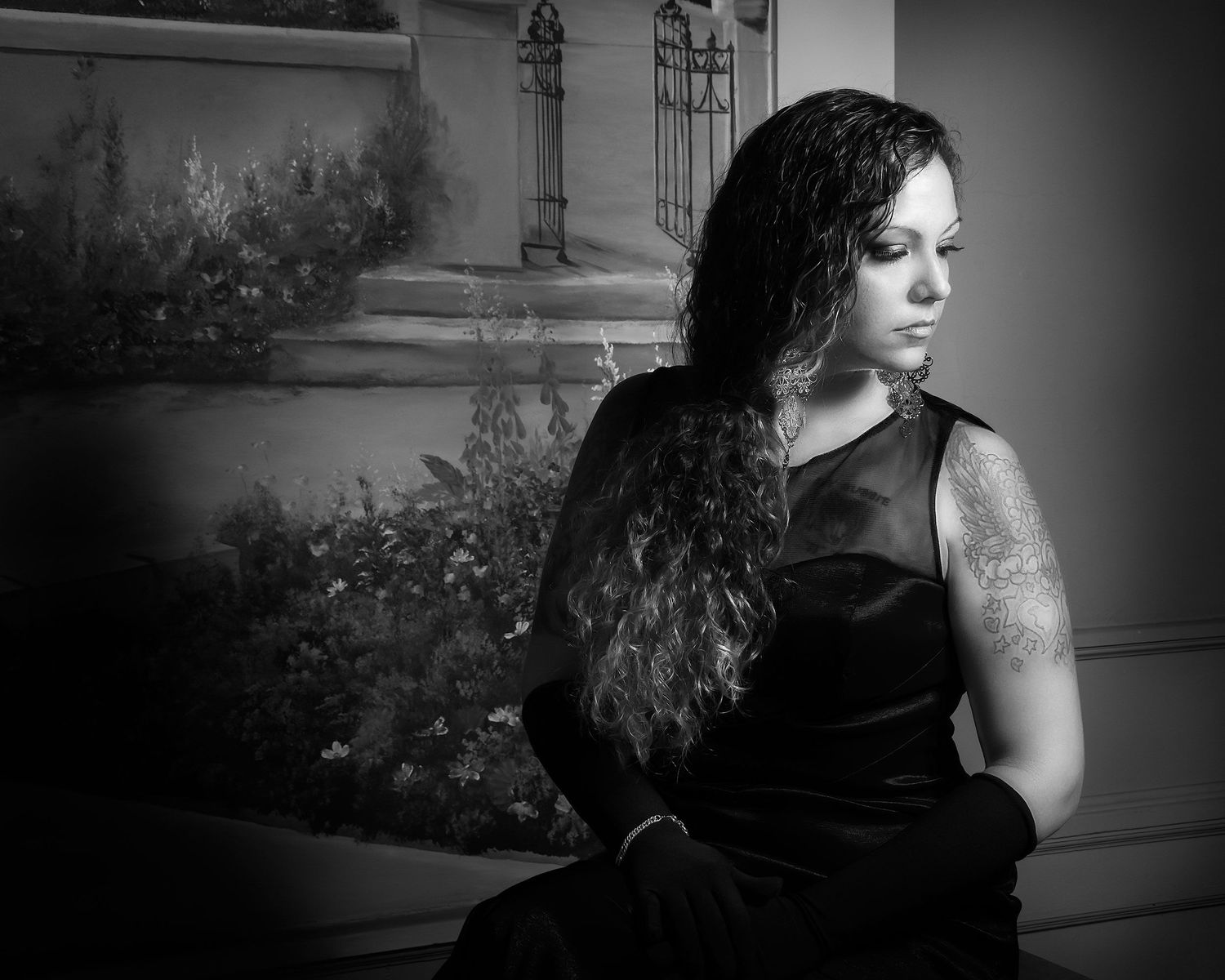 Black and white portrait of a woman in black dress with tattoo, sitting pensively against decorative backdrop.