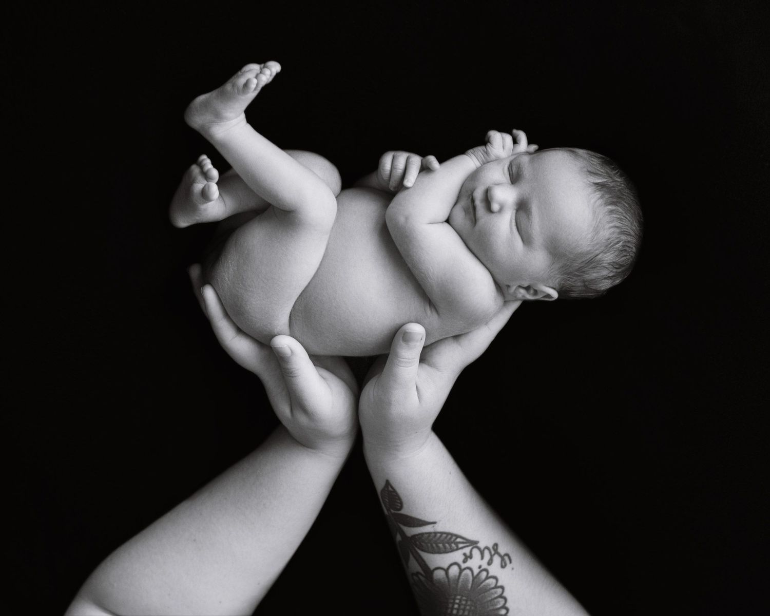 Black and white photo of a newborn baby cradled in tattooed hands against a dark background.
