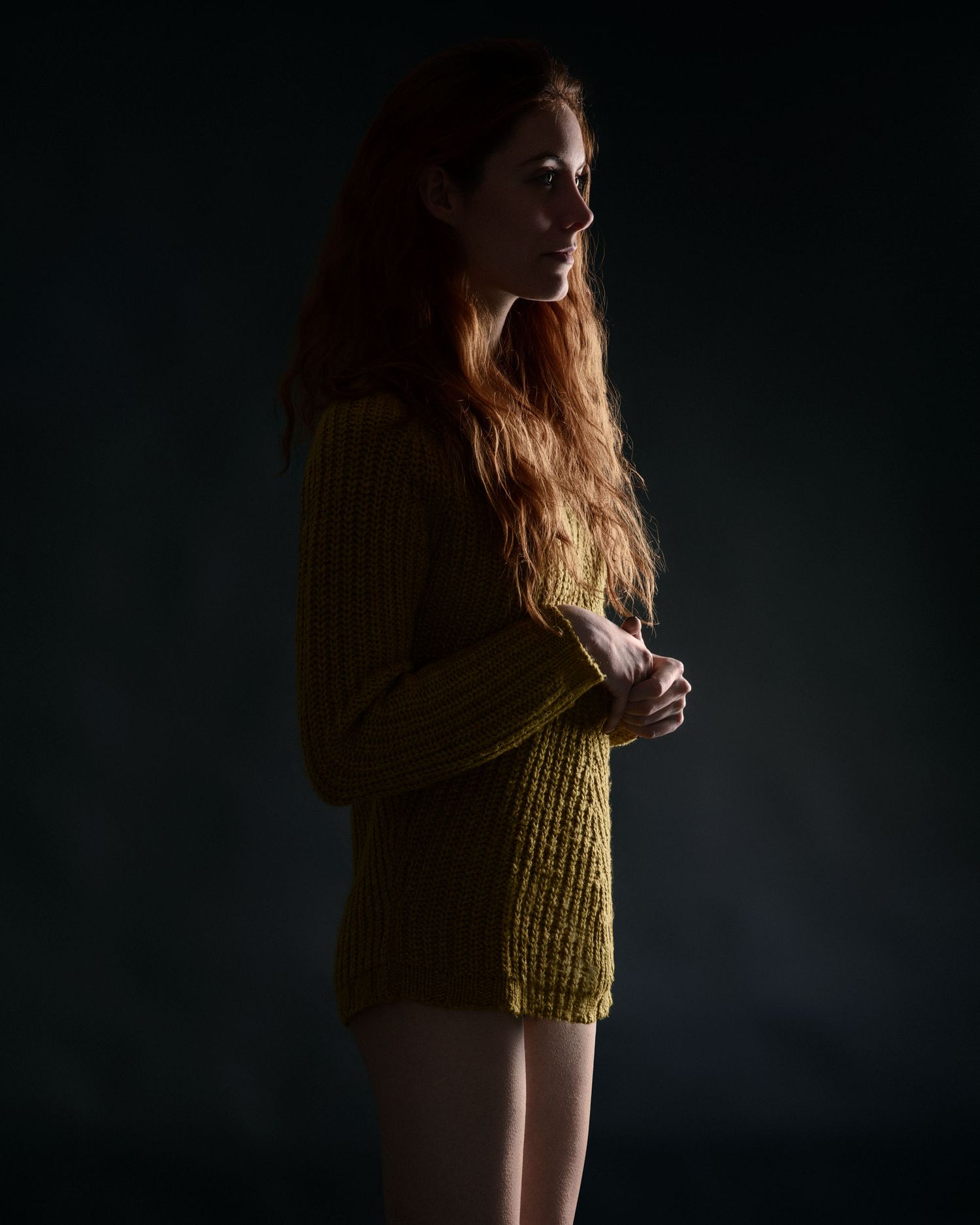 A young woman with long red hair wearing an oversized yellow knit sweater poses in profile against a dark background.