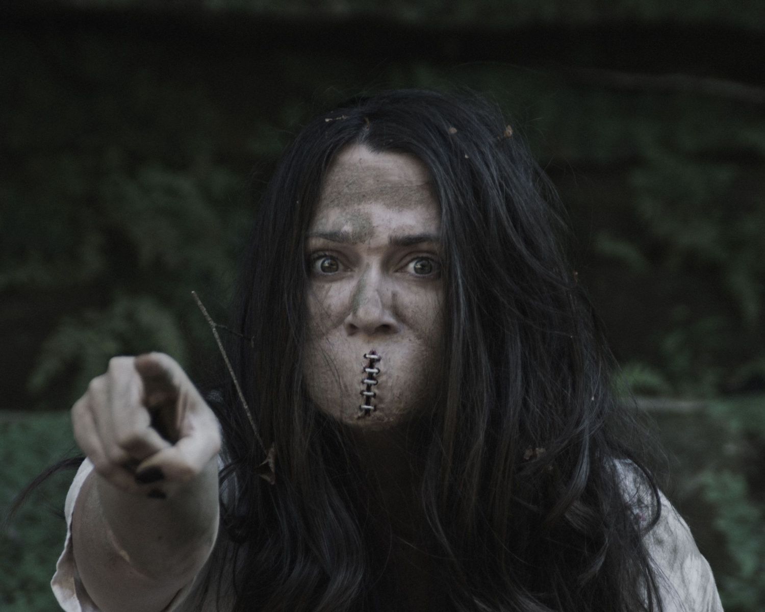 Creepy horror character with long black hair and zipper mouth frantically pointing outdoors.