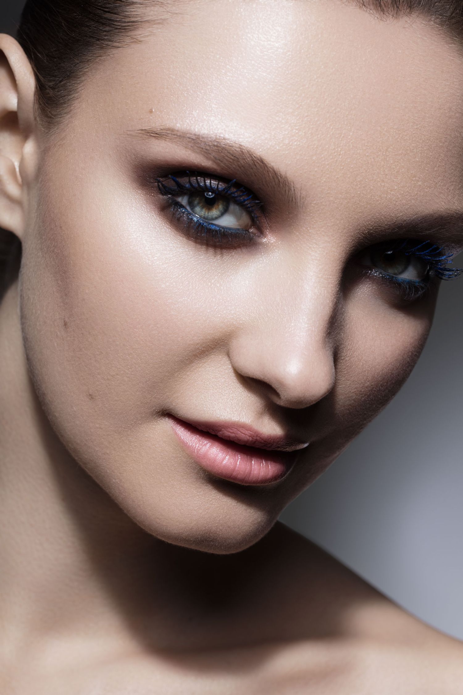 Close-up portrait of a woman with smoky eye makeup featuring blue mascara and defined brows on a neutral background.
