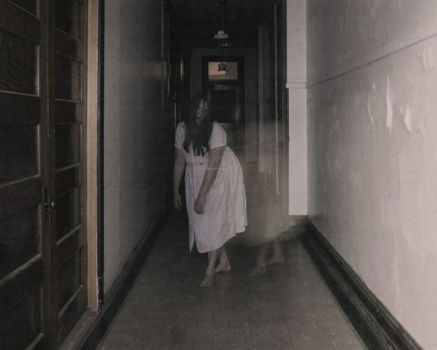 Eerie figure in white dress walks dimly lit hallway in ghostly horror scene.