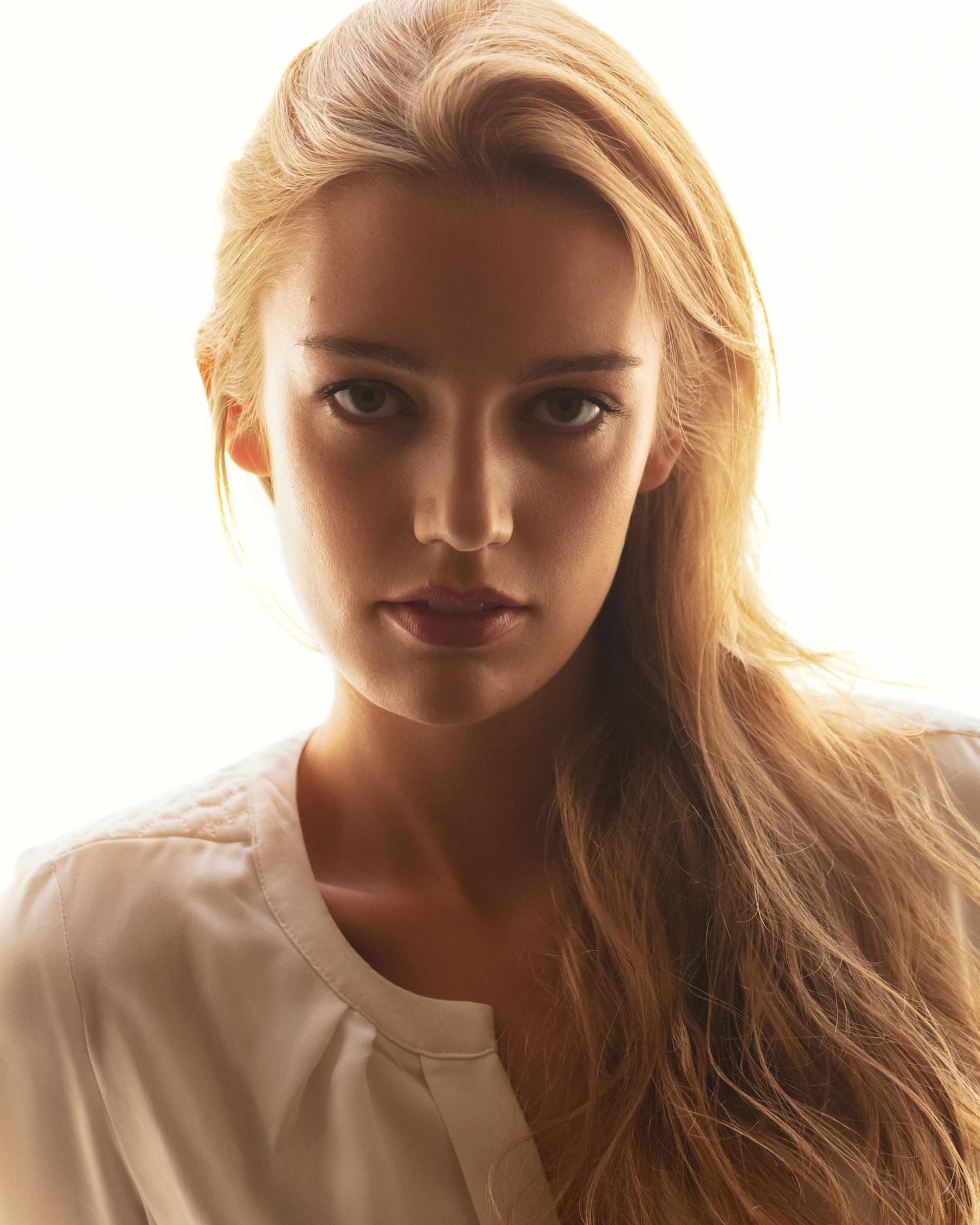 Young blonde woman with long hair wearing a white blouse, captured in a bright, softly lit portrait photo.